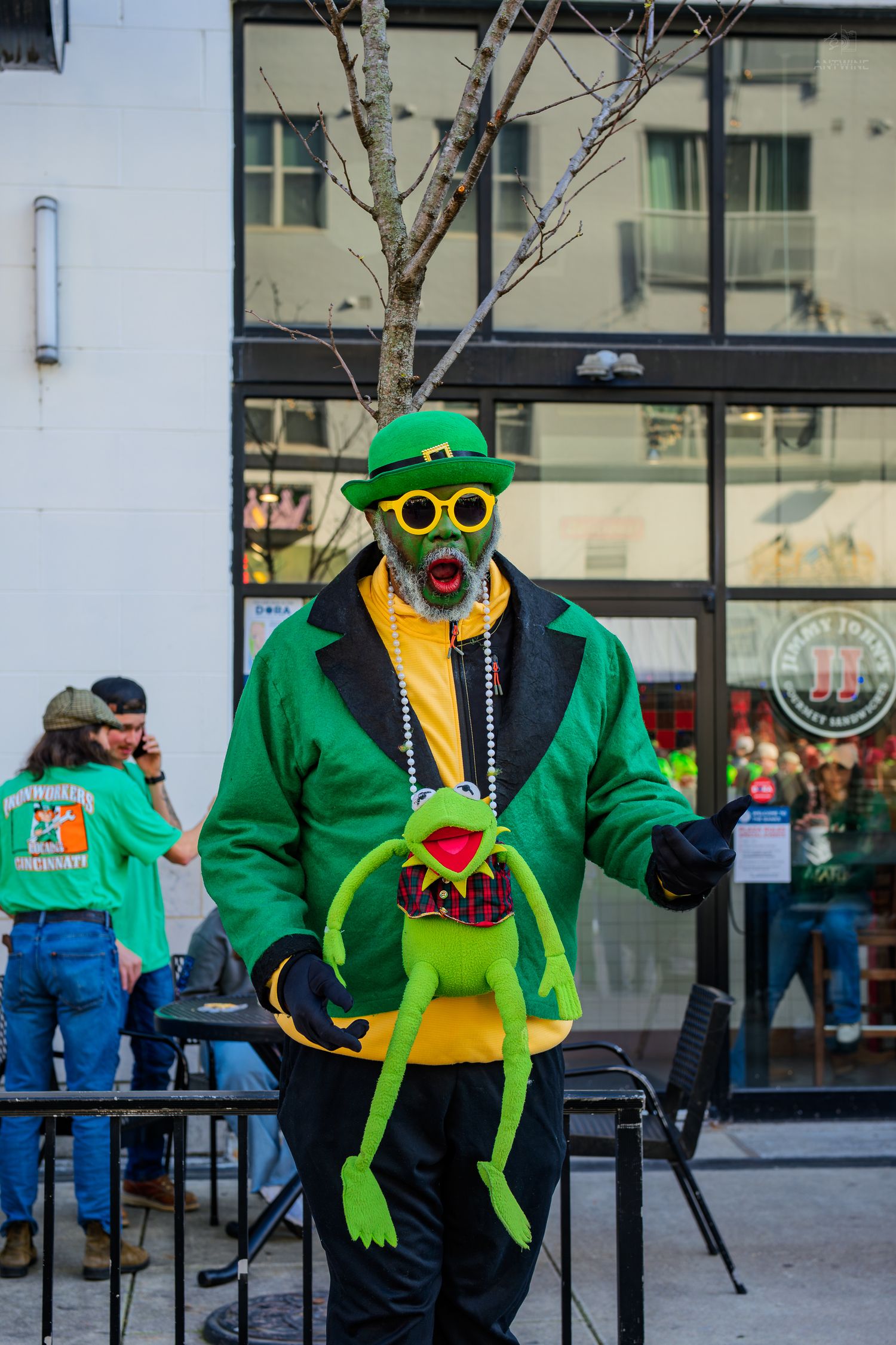 Man in green leprechaun costume holding Kermit the Frog doll at St. Patrick's Day parade celebration.