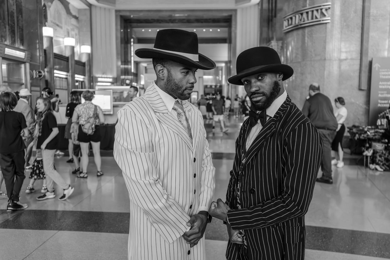 Two stylish men in pinstripe zoot suits and wide-brim hats pose in a grand train station.