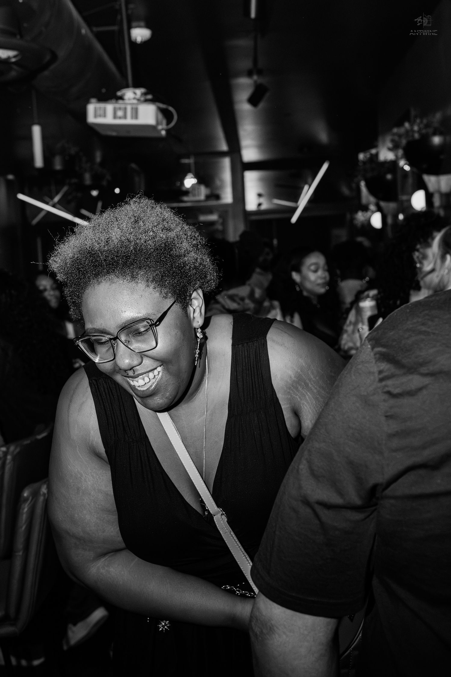 A smiling Black woman wearing glasses and a black dress laughs joyfully at a dimly lit social gathering.