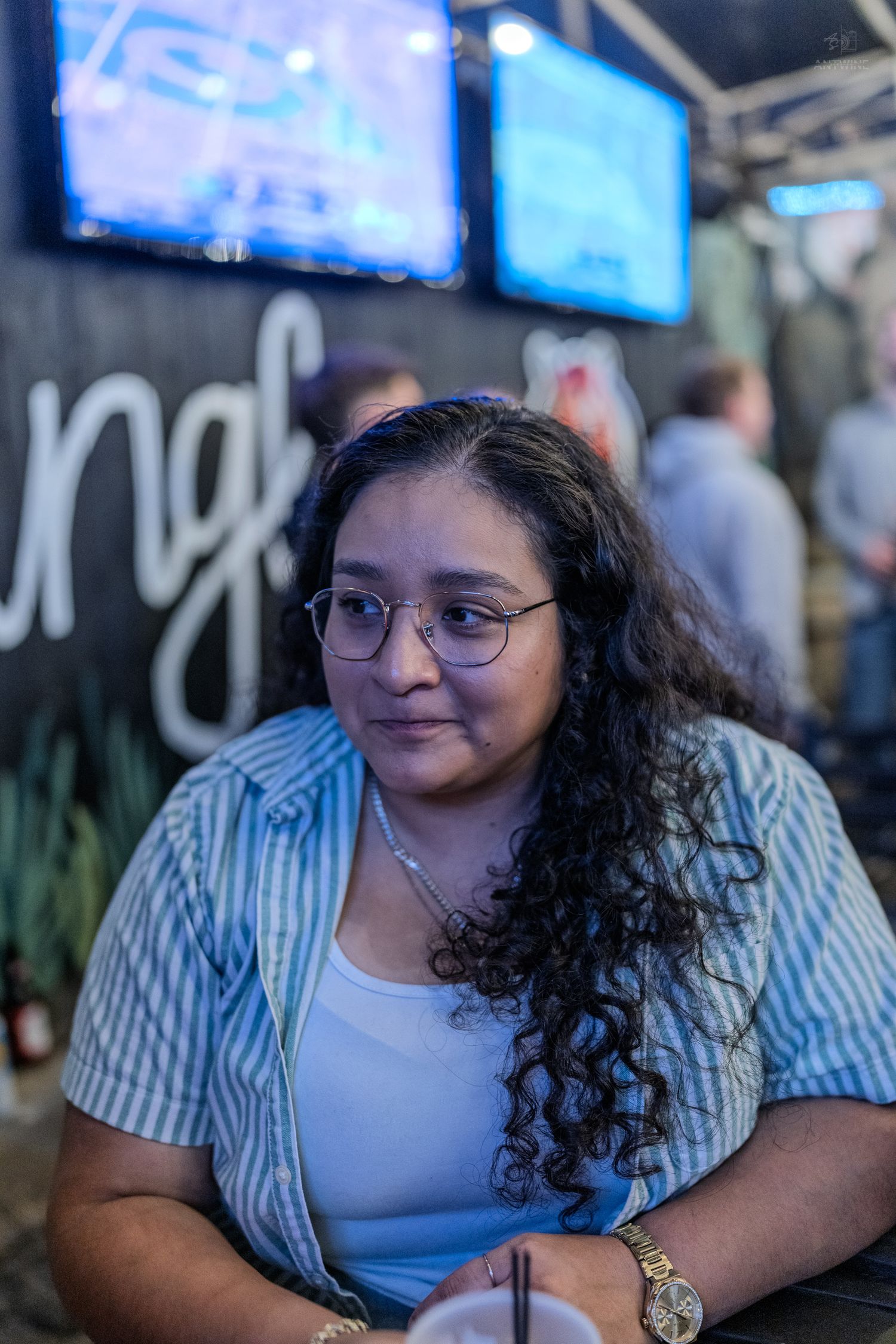 Young woman with curly hair and glasses smiling at a bar with TV screens in the background.