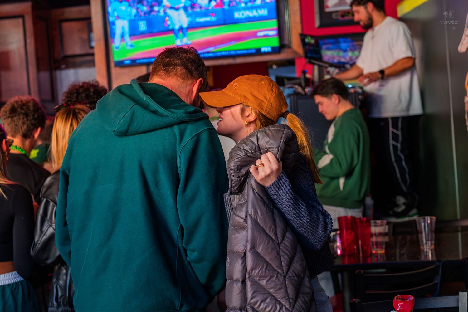 Couple sharing an intimate moment at a sports bar with a football game on TV in the background.