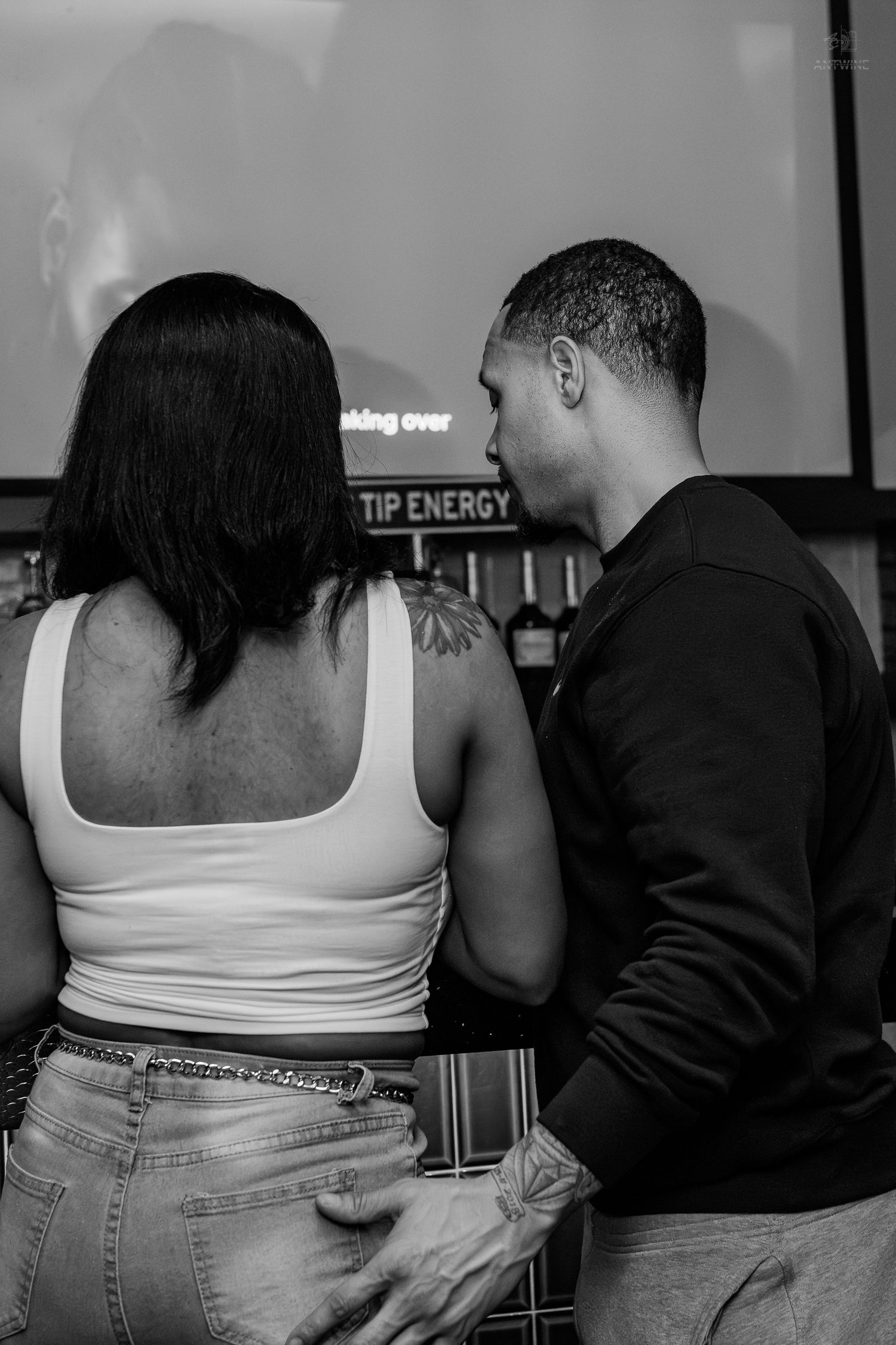 Black and white photo of a couple standing together at a bar, woman with tattoo wearing white tank top and jeans.