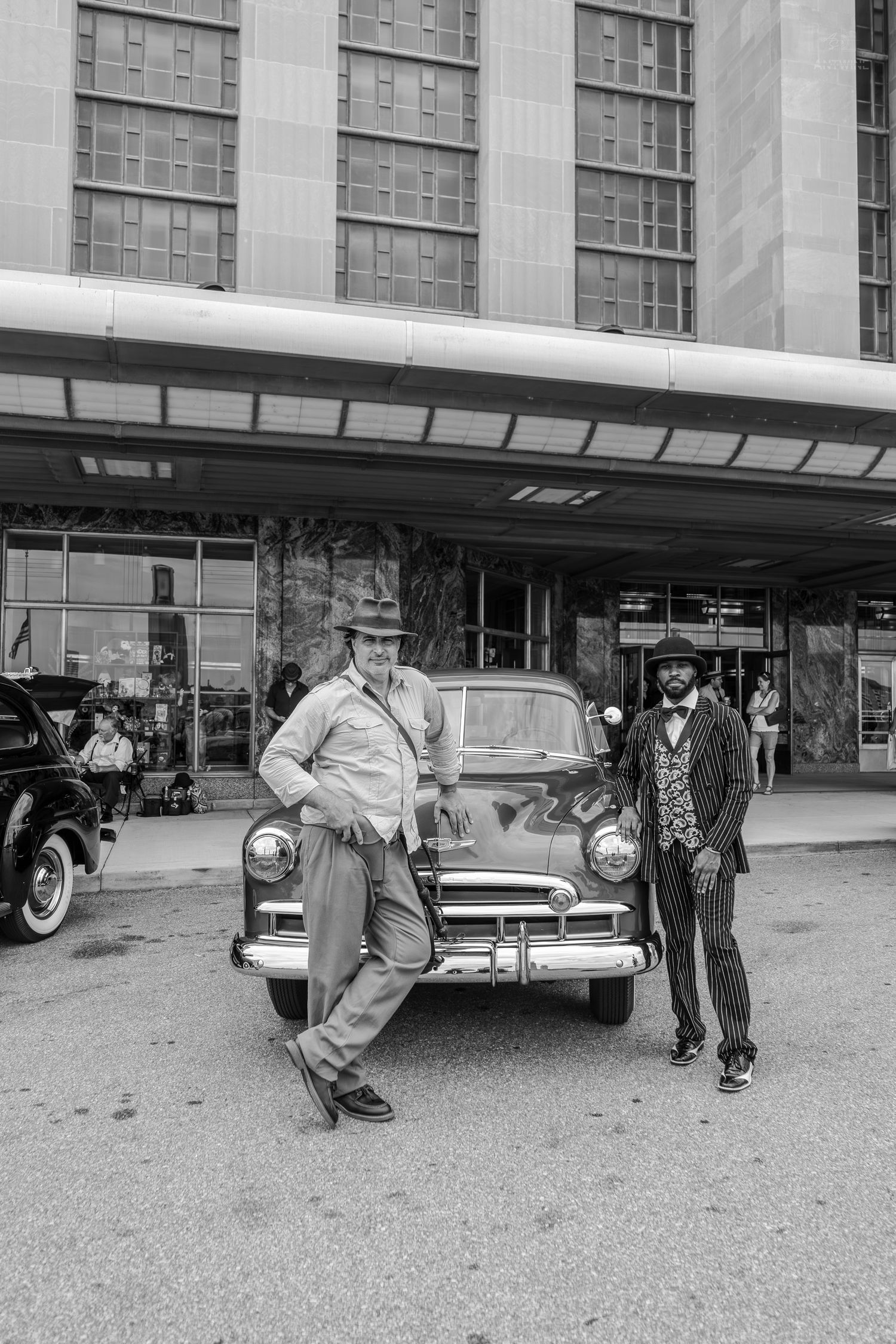 Two men in vintage attire pose beside a classic 1950s car outside a historic building in black and white.