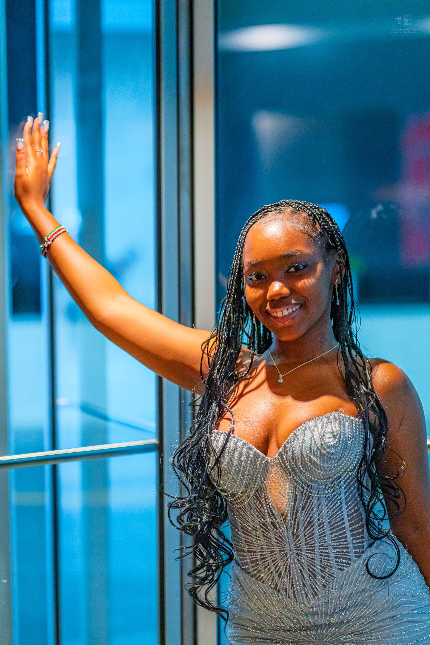 Young woman in a silver sequin gown with braids waving and smiling near a glass wall.
