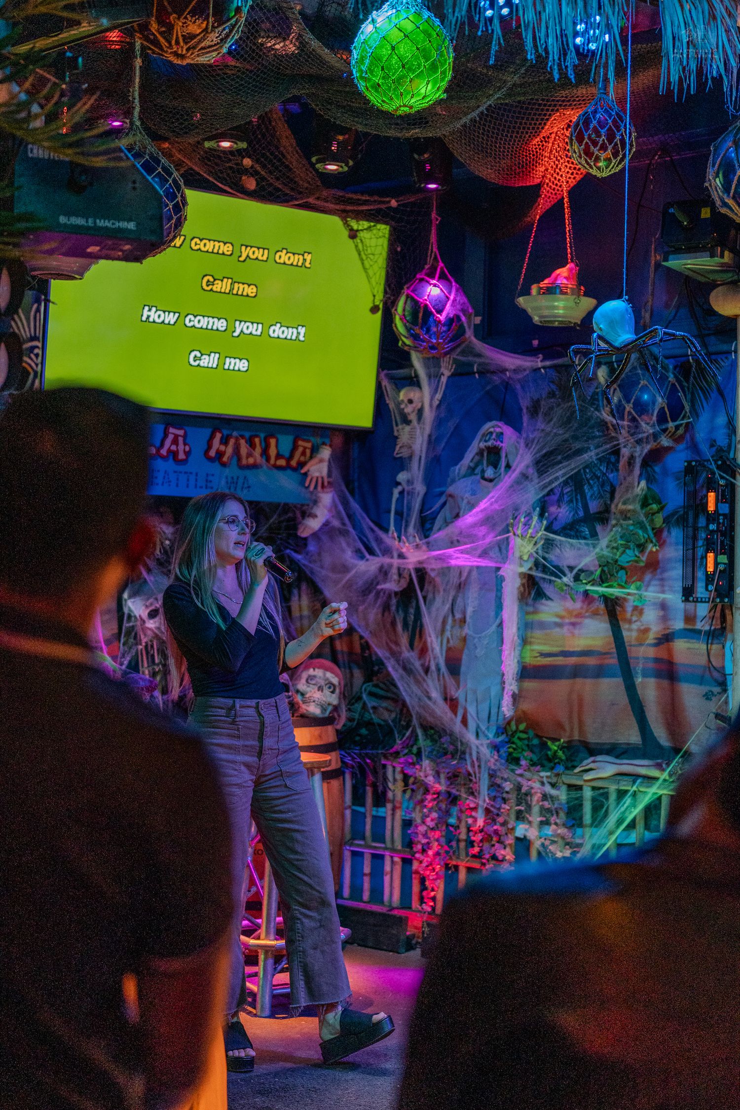 Woman singing karaoke at a Halloween-decorated bar with colorful lights and spooky cobweb decorations.