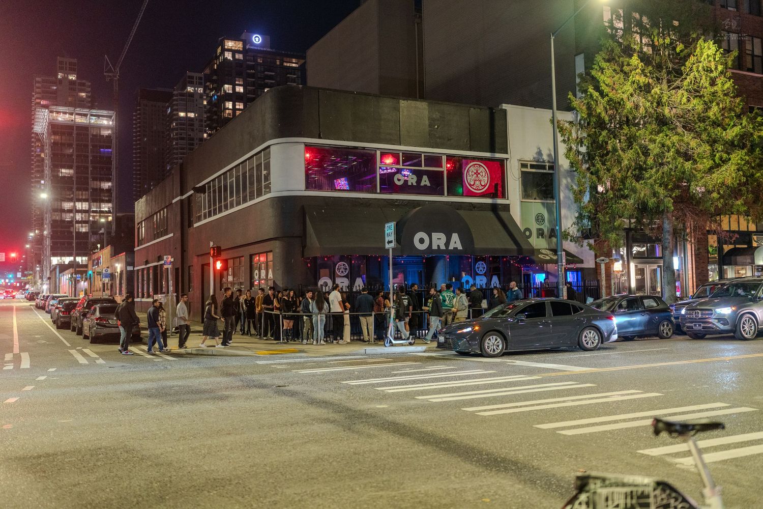 Crowd lines up outside ORA nightclub at night on a busy urban street corner with city buildings lit up.