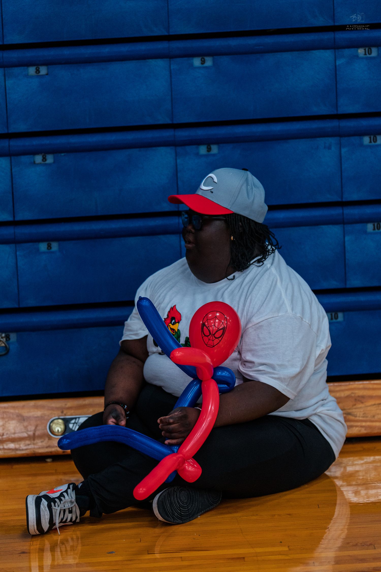 Person sitting on gym floor holding red and blue balloon animals, wearing white tee and gray cap.