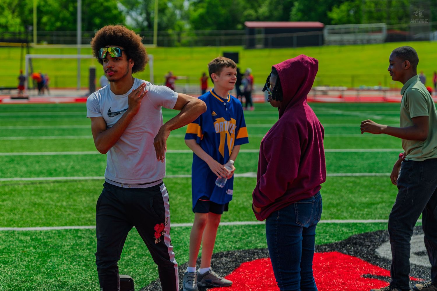 Young athletes converse on a red and green football field during an outdoor sports event on a sunny day.