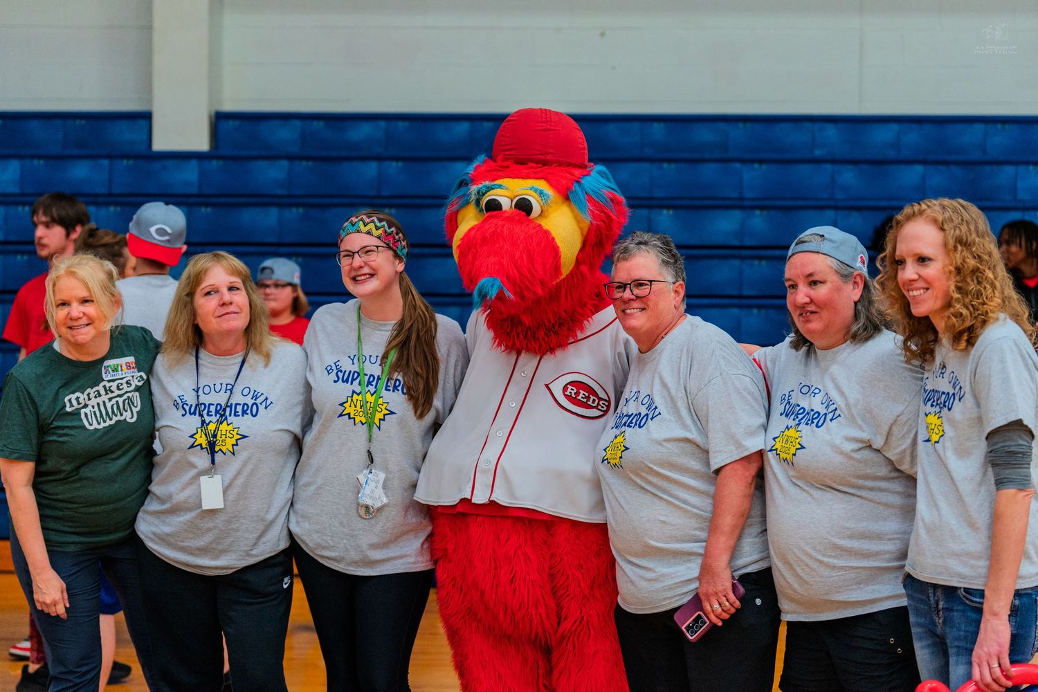 Group of women in matching gray t-shirts posing with a red bird mascot in a gymnasium.