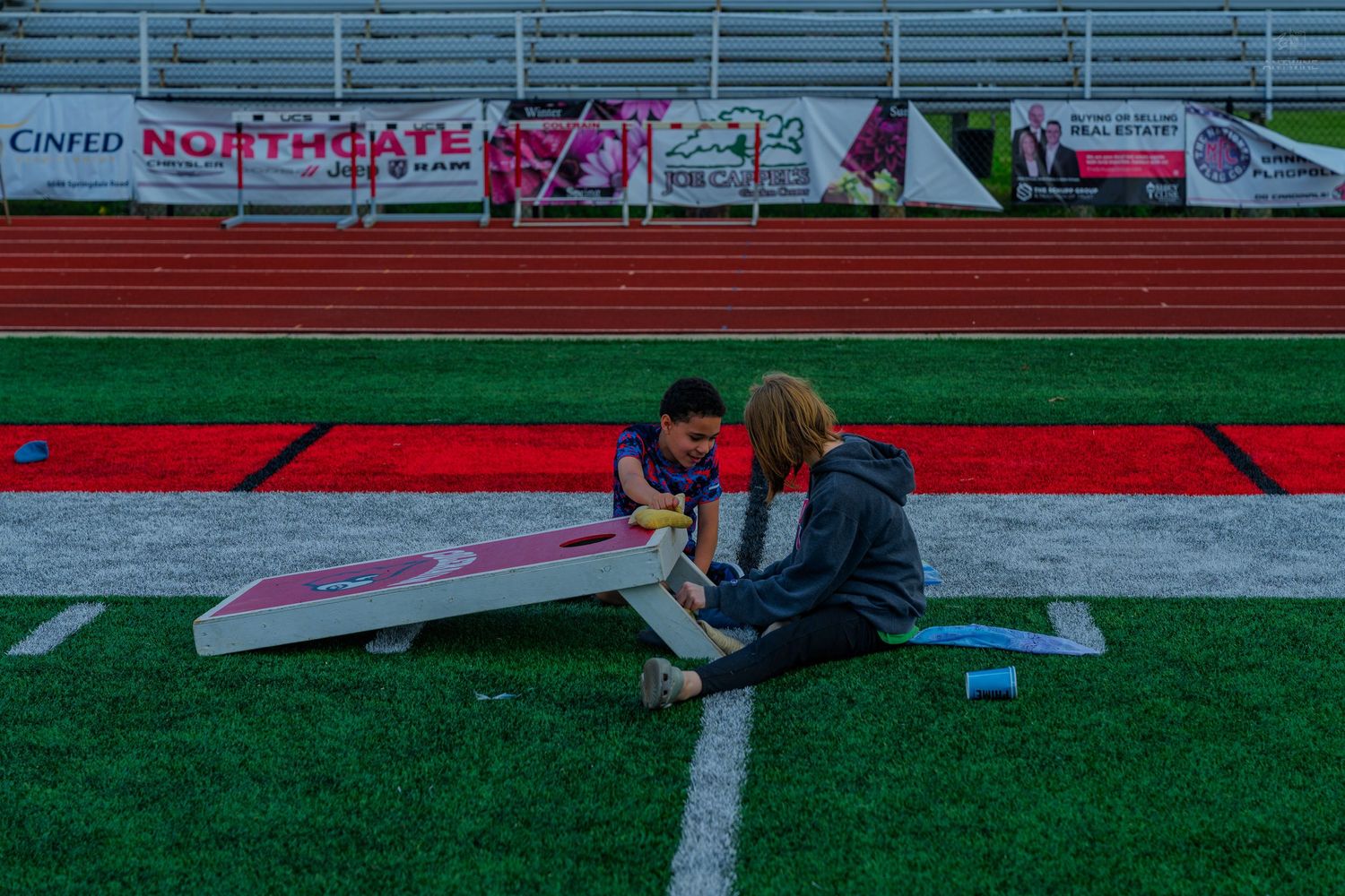 Two people painting a cornhole board on a football field near red track and stadium signage.