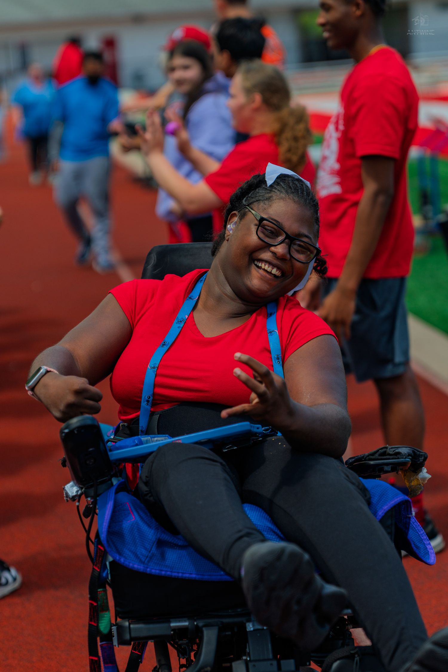 Smiling young woman in wheelchair wearing red shirt and glasses, making peace sign at outdoor track event.