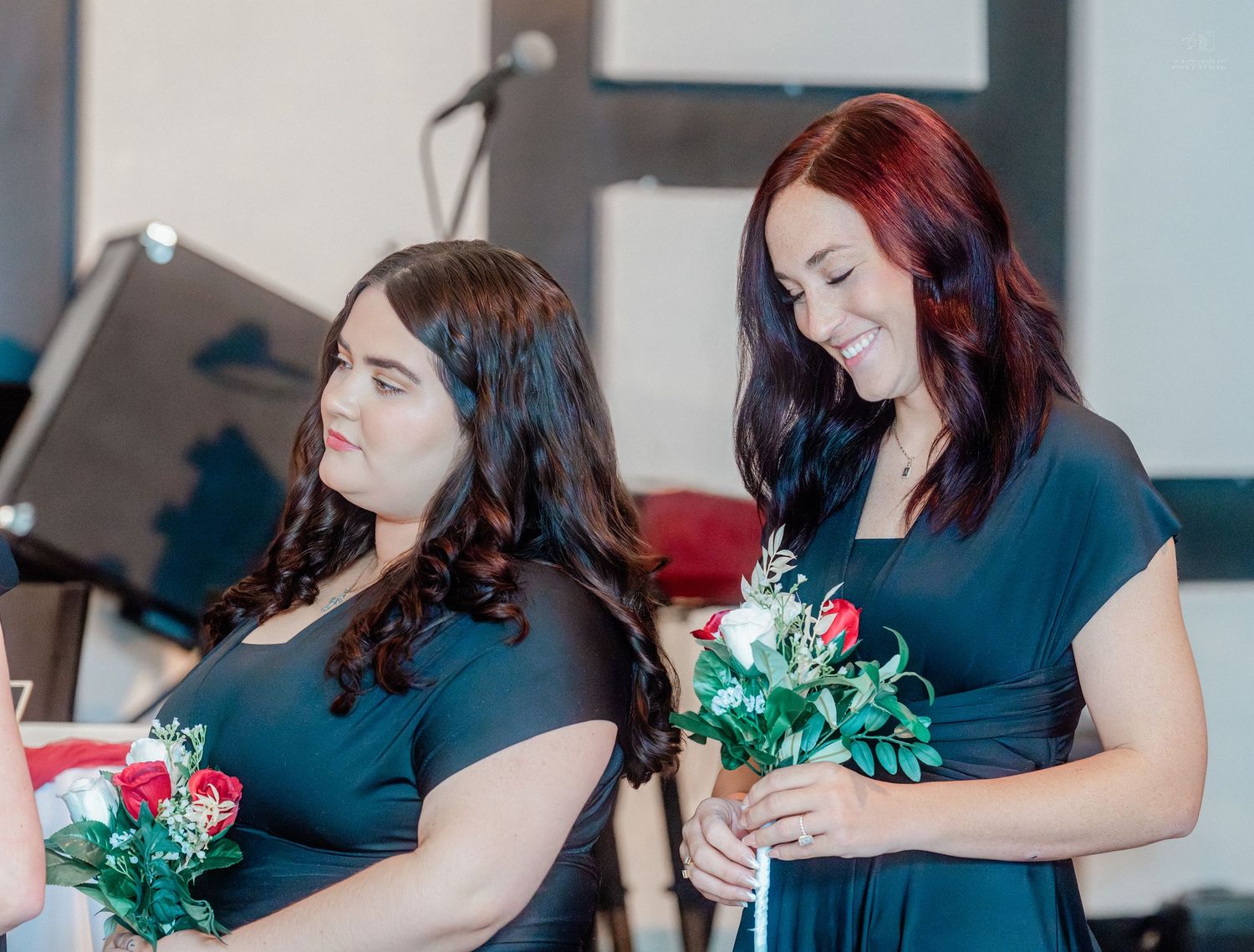 Two bridesmaids in navy dresses holding floral bouquets with red roses smile during a wedding ceremony.