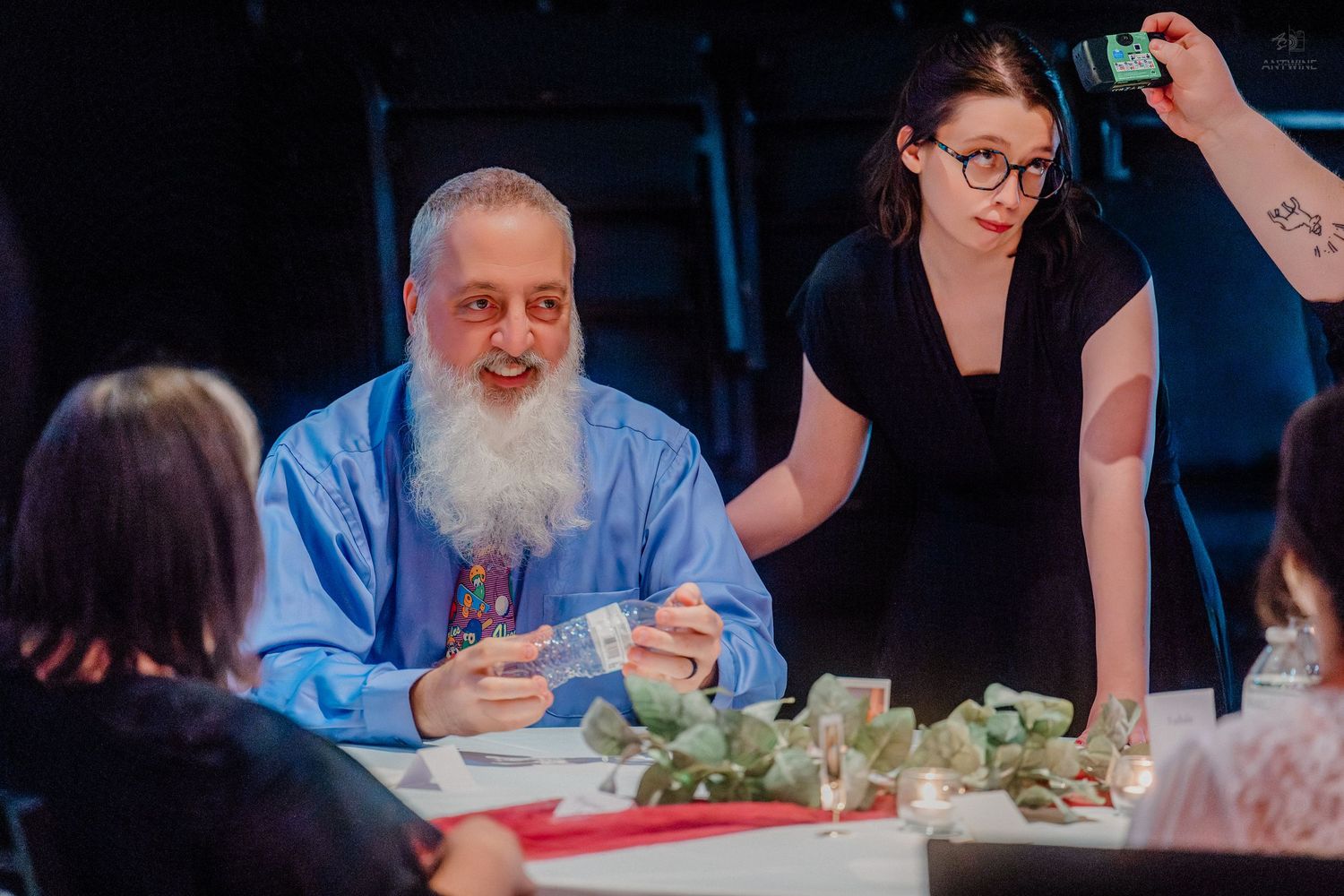 Elderly bearded man in blue shirt examines greenery at table while young woman with glasses assists at event.