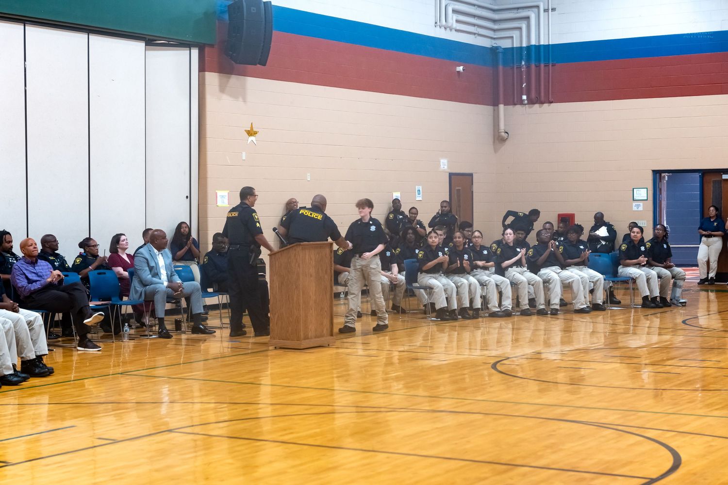 Students and officers gather in a school gymnasium for a formal ceremony, seated in rows on a polished hardwood floor.