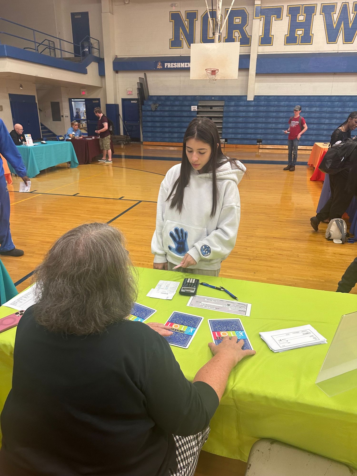 Student reviews materials at a bright green table during a school fair inside a Northwest high school gymnasium.