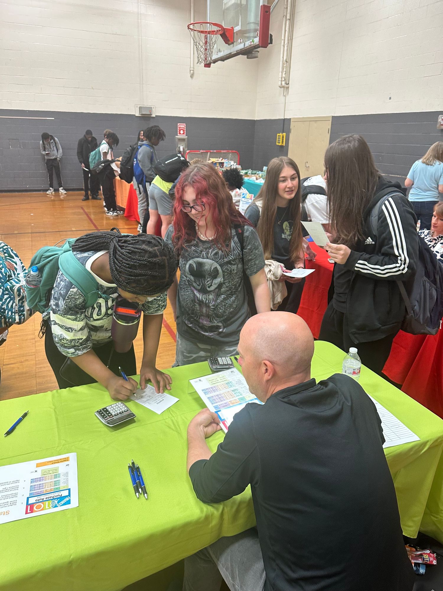 Students gather around a green-draped table interacting with a seated representative at a school gymnasium fair.