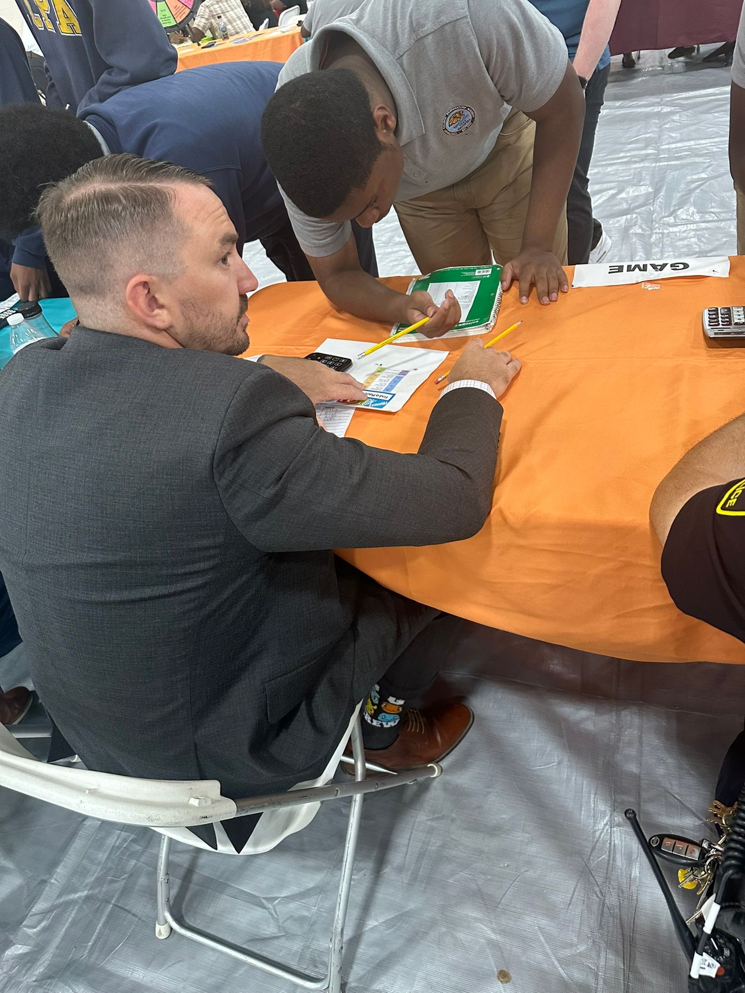 Man in gray suit signing documents at an orange table during a professional event or job fair.