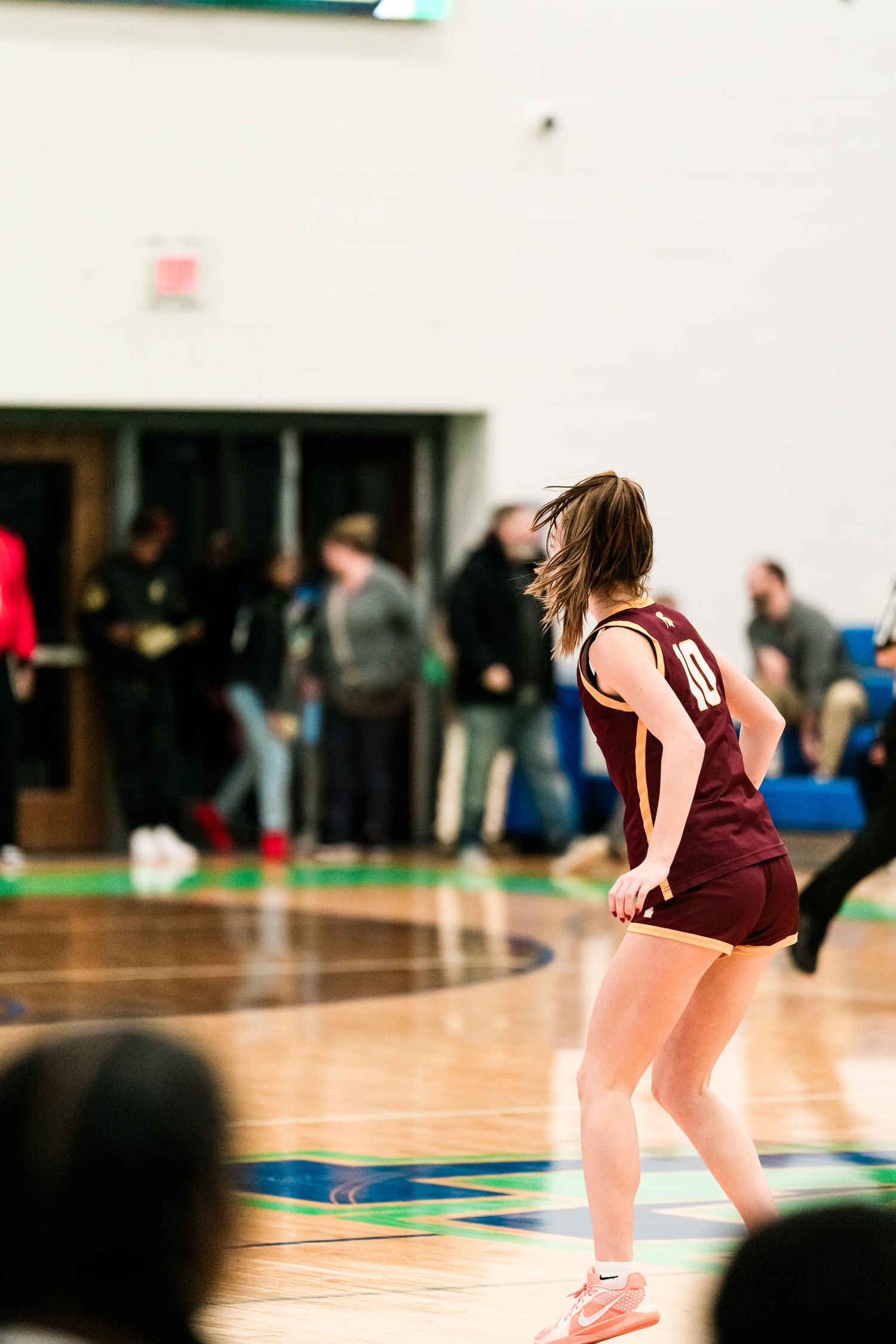 Female high school basketball player in maroon uniform crouches in defensive stance on a hardwood court during a game.