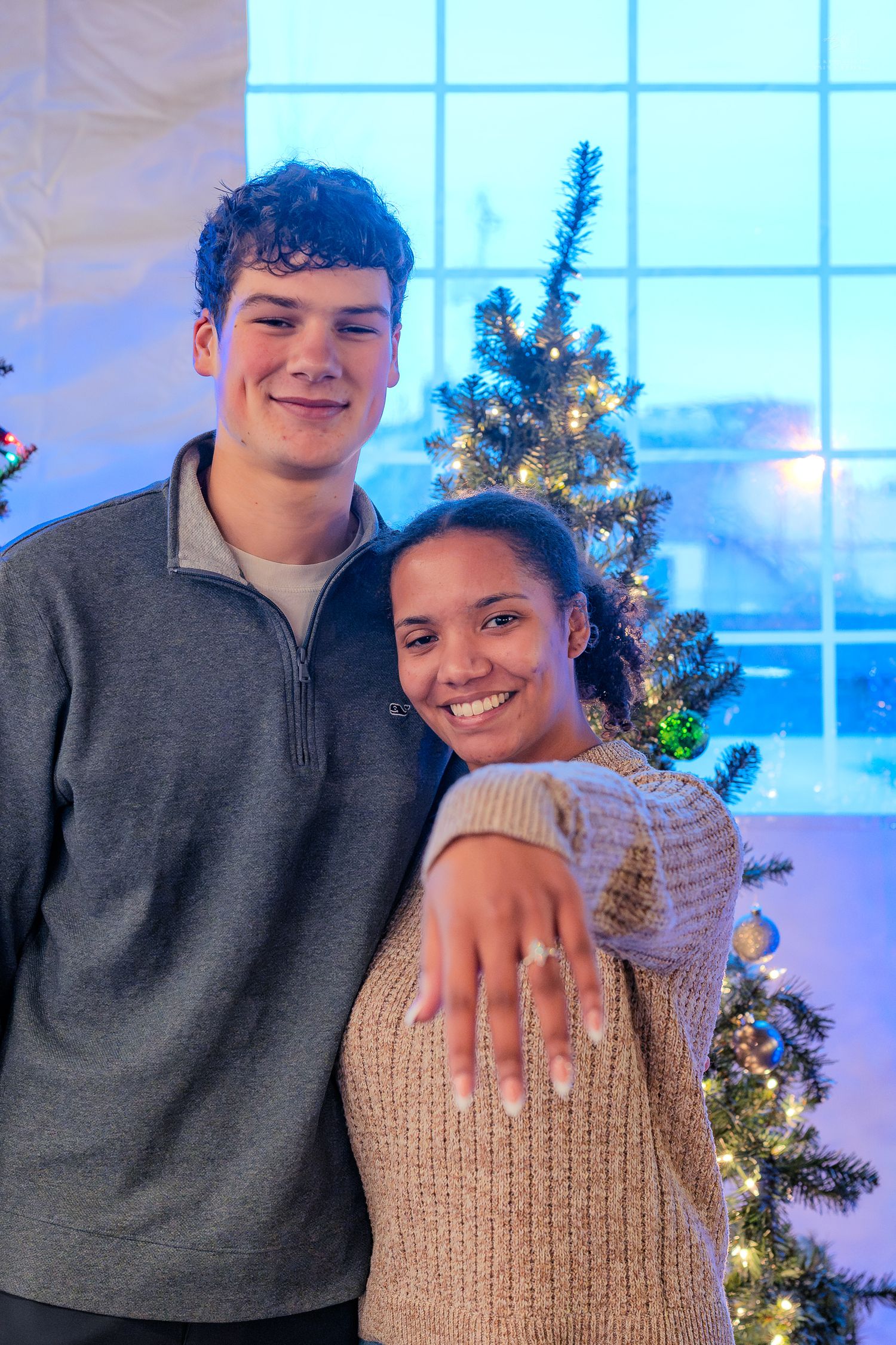 Young couple poses by Christmas tree as woman shows off engagement ring.