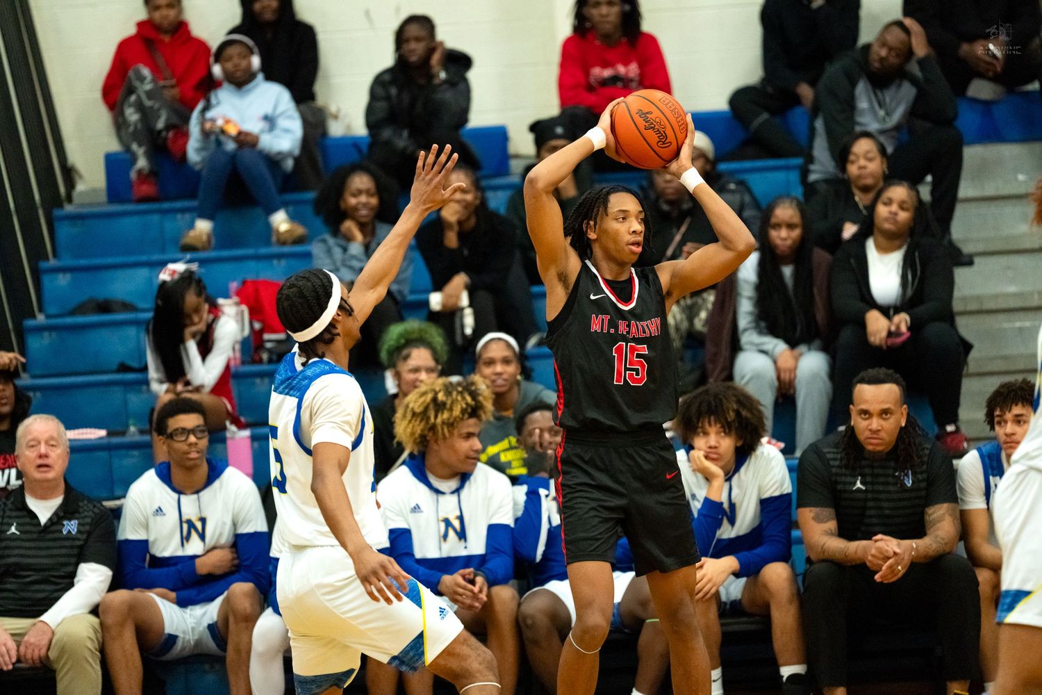Player #15 in black uniform shoots basketball over defenders in high school game.