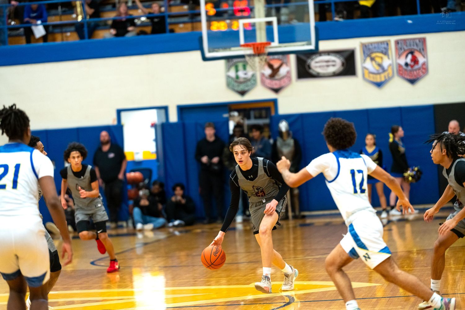 Basketball player in dark uniform dribbles between defenders during high school indoor basketball game.