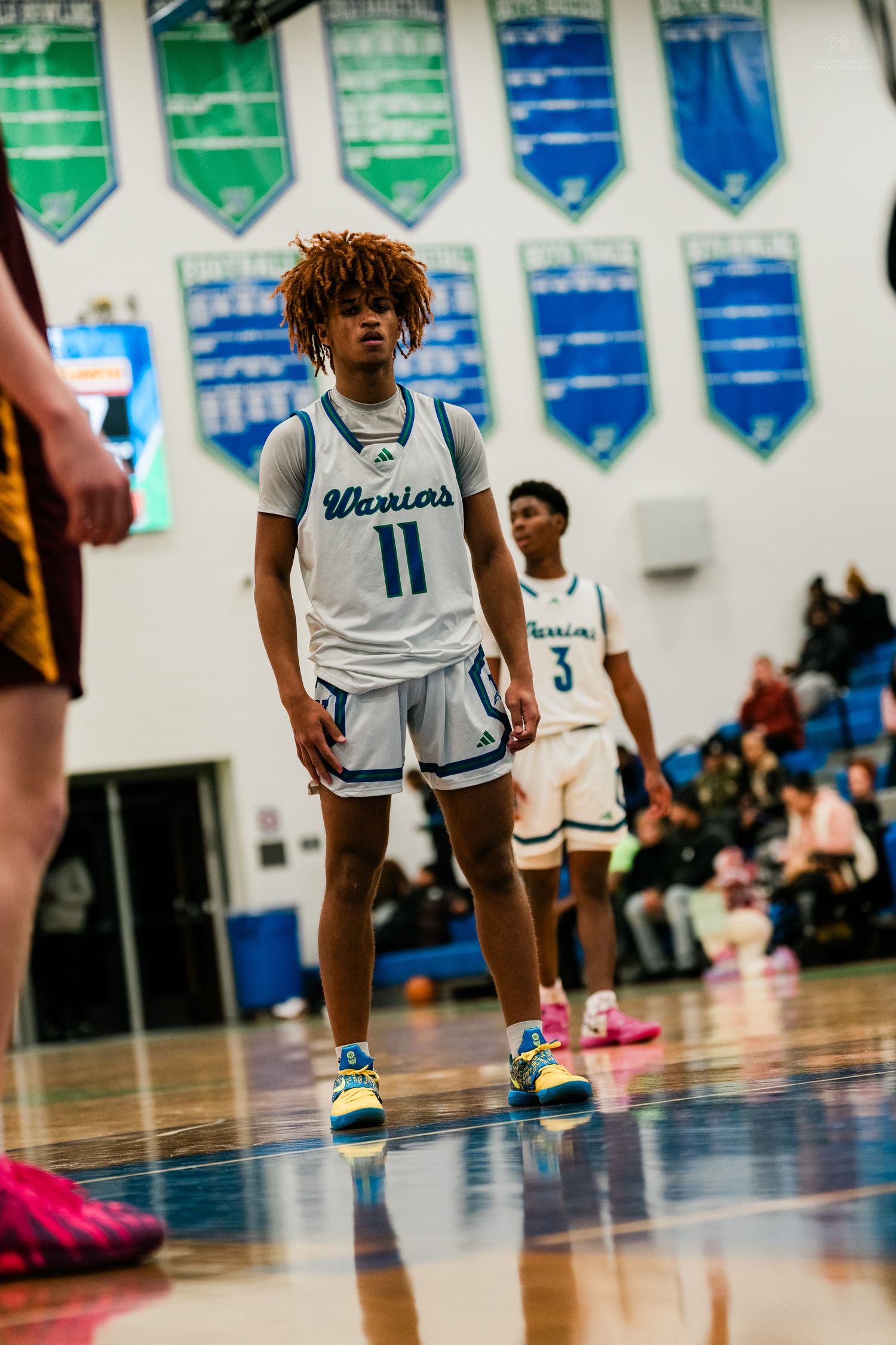 Warriors basketball player number 11 stands on the court during a high school game with teammate number 3 behind him.
