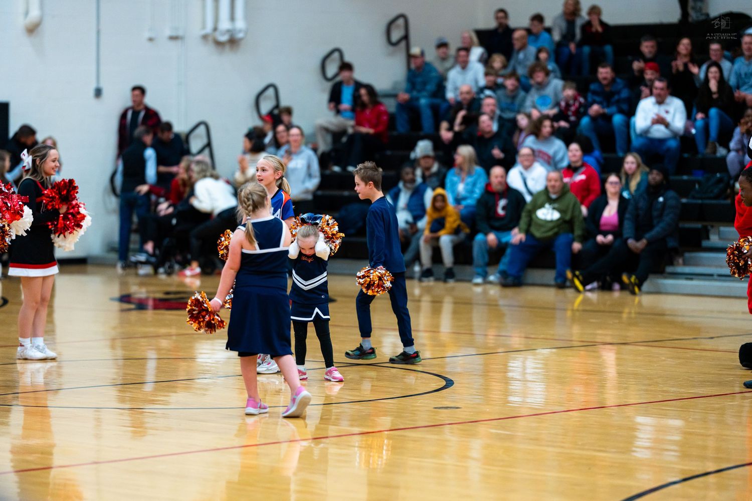 Young cheerleaders and children in navy uniforms stand on basketball court before a packed gymnasium crowd.