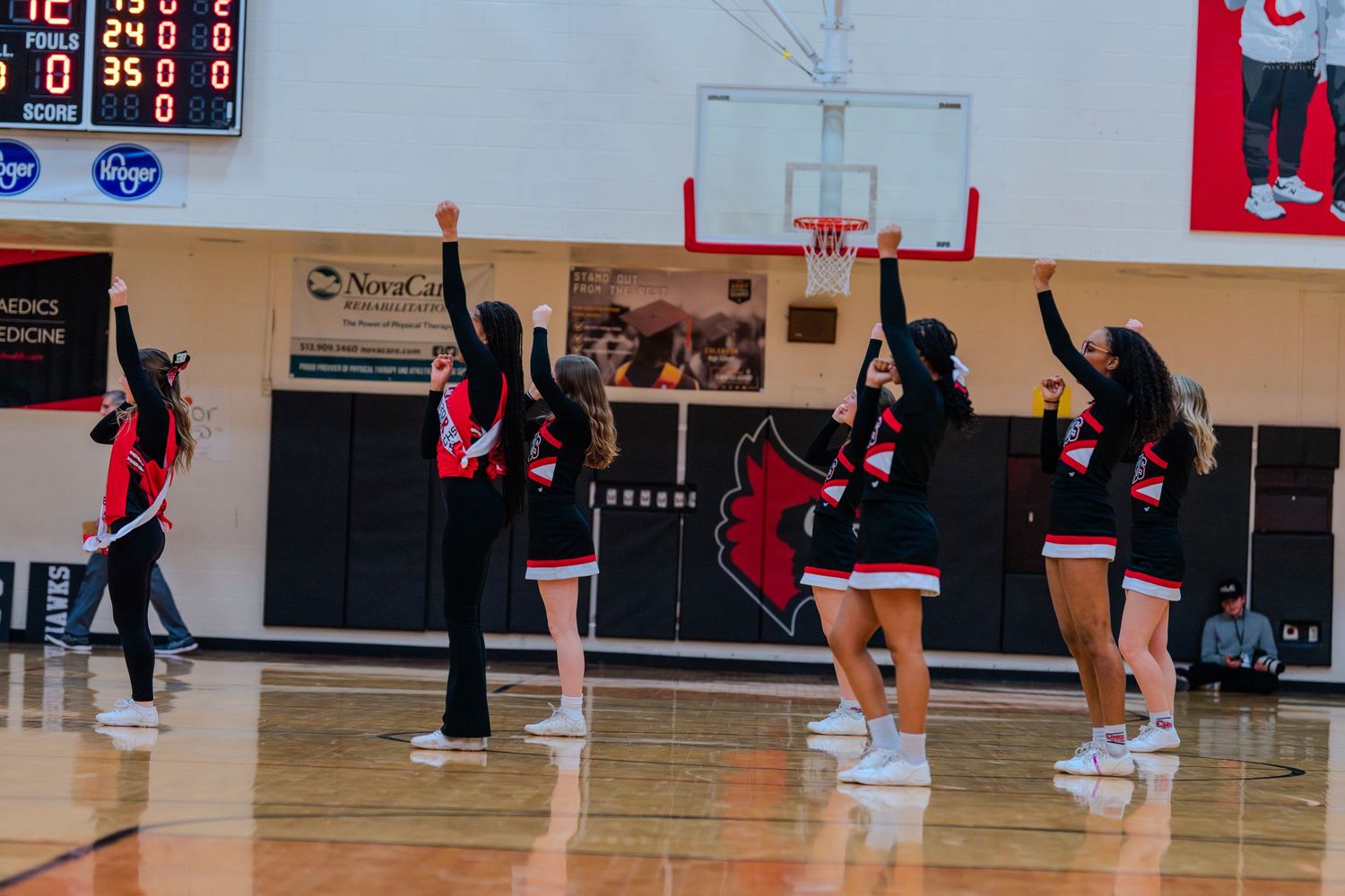 High school cheerleaders in red and black uniforms raise their arms during an energetic sideline performance in a gym.