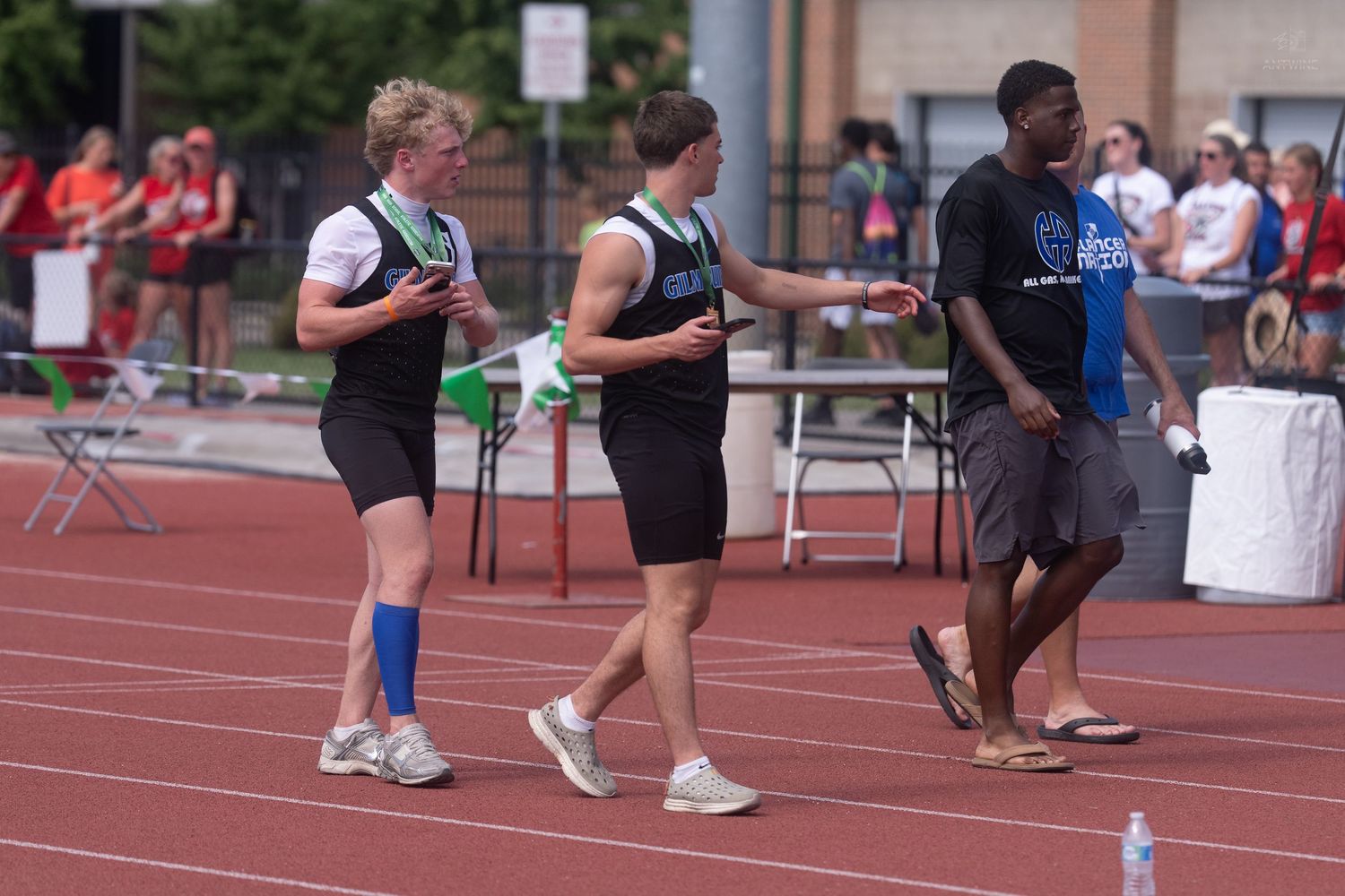 Three male track athletes in green uniforms interact on a red track during an outdoor relay competition.