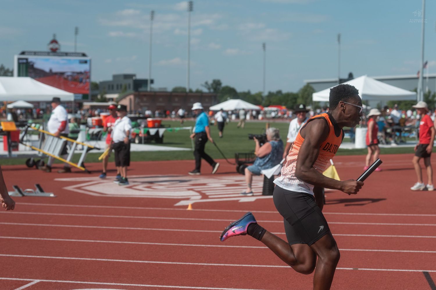 Male sprinter runs with a baton on a red track during an outdoor relay race at a track and field competition.
