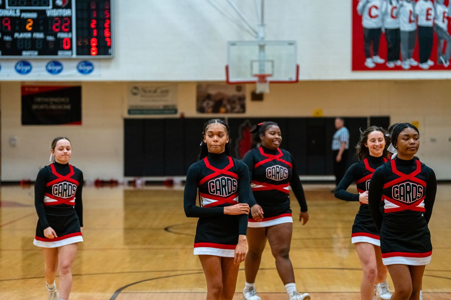 Five cheerleaders in black and red CHHS uniforms walk onto a basketball court during a game.