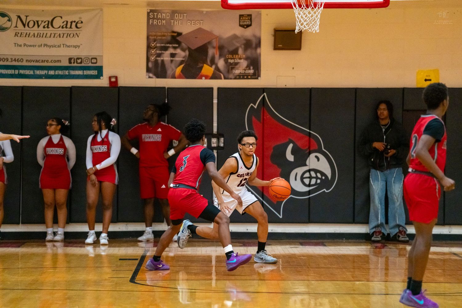 Basketball player in white dribbles past red-uniformed defender near Cardinals logo court during high school game.
