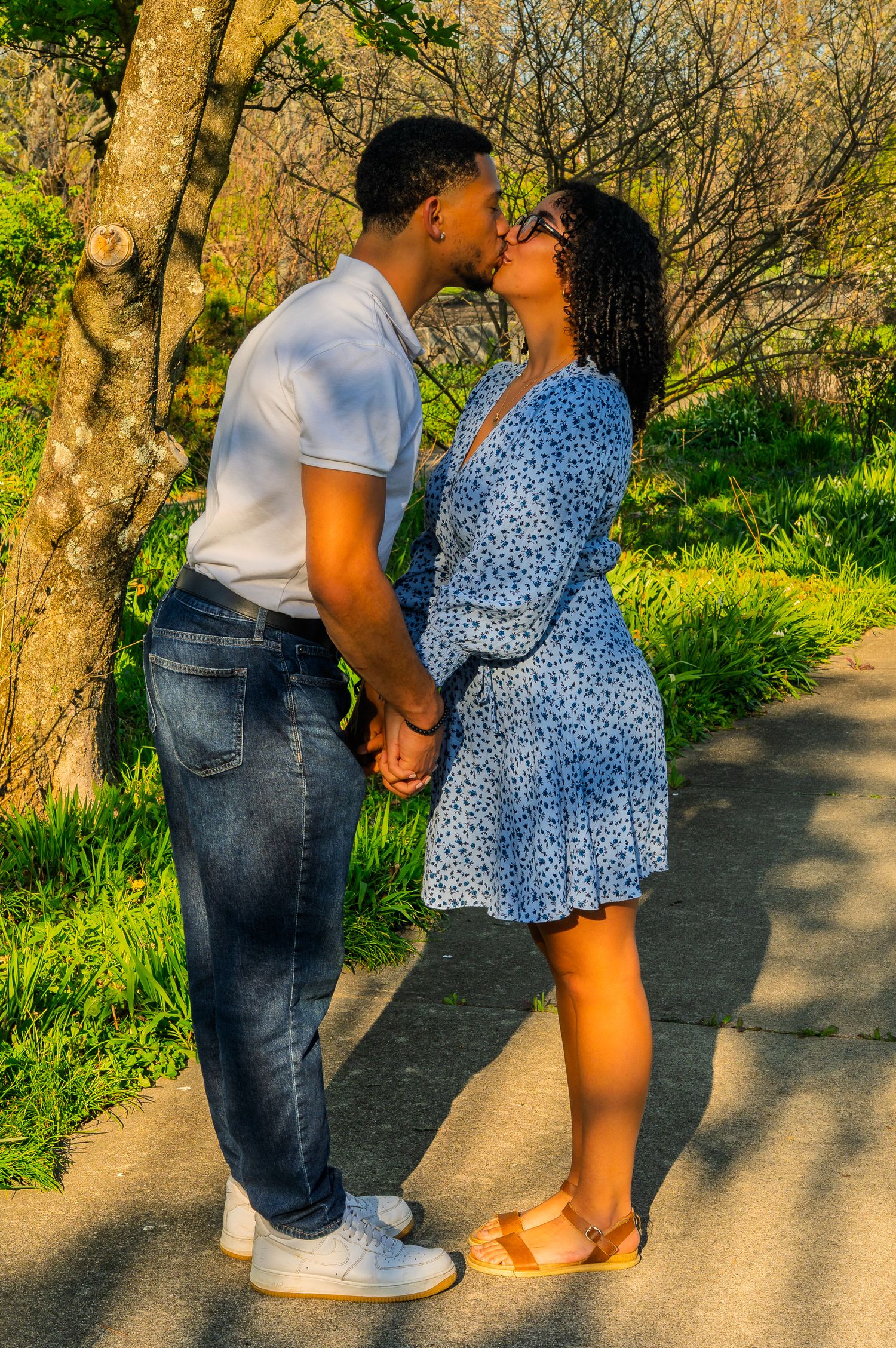 Couple kissing while holding hands on a sunlit park path surrounded by greenery.