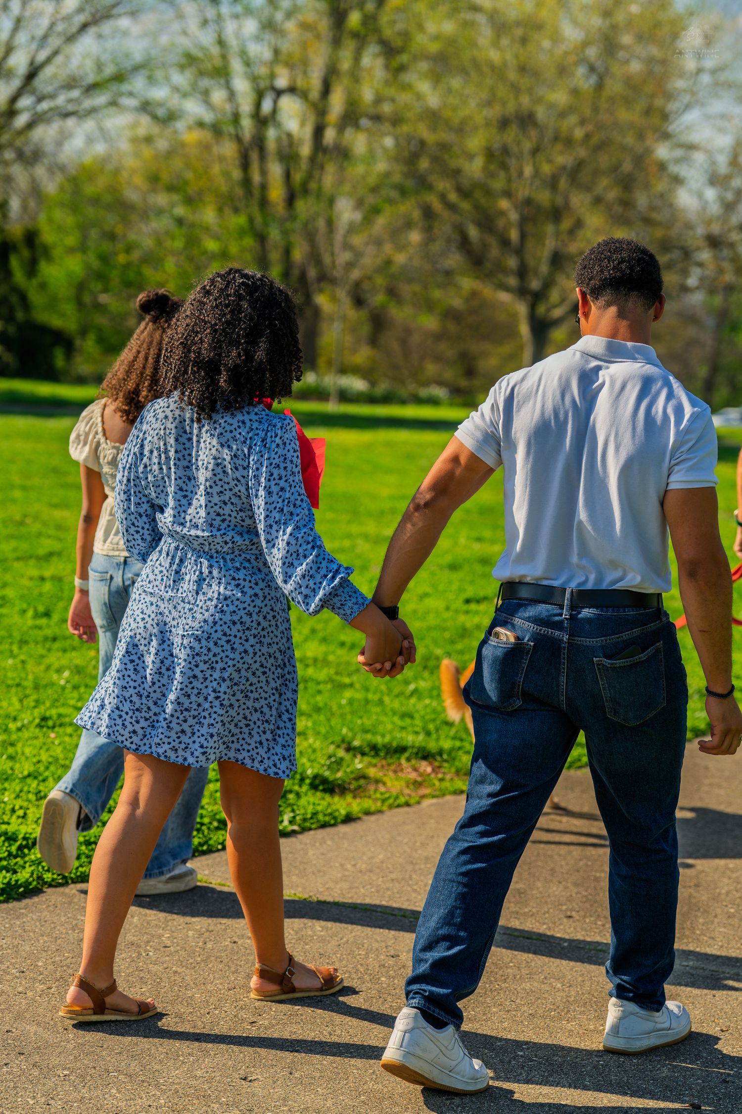Couple holding hands walking on a park path, woman in blue floral dress and man in white shirt and jeans.