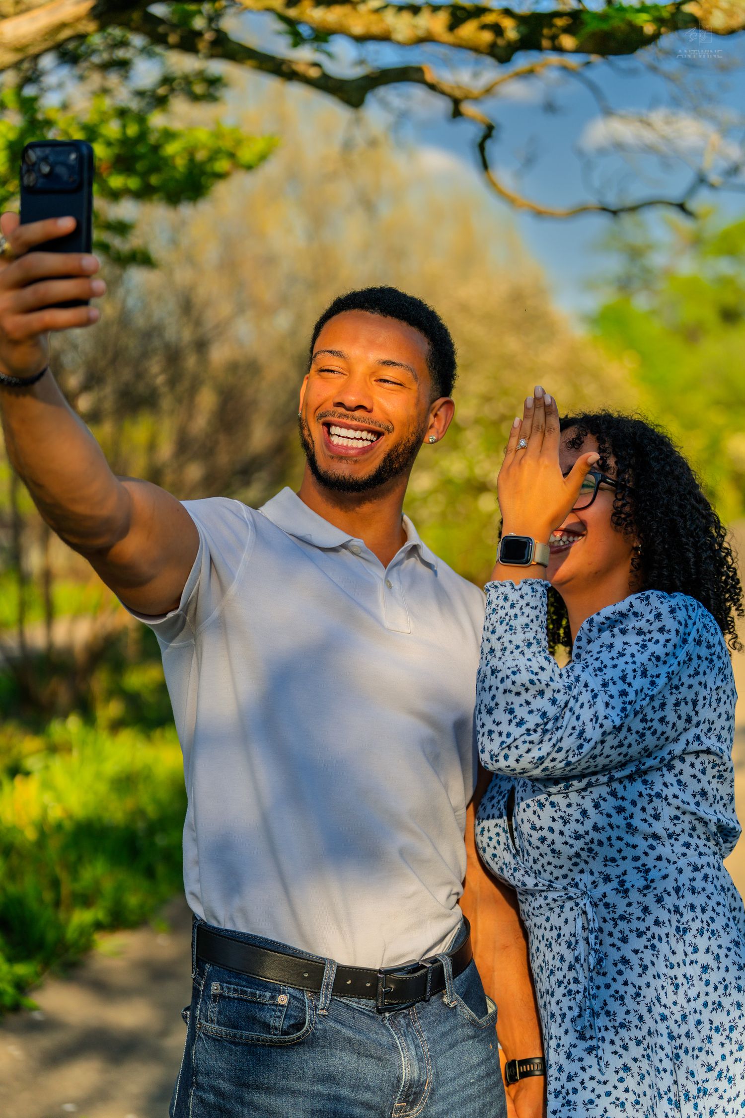 A joyful couple takes a selfie outdoors, woman covering mouth in surprise, man smiling broadly holding phone up.
