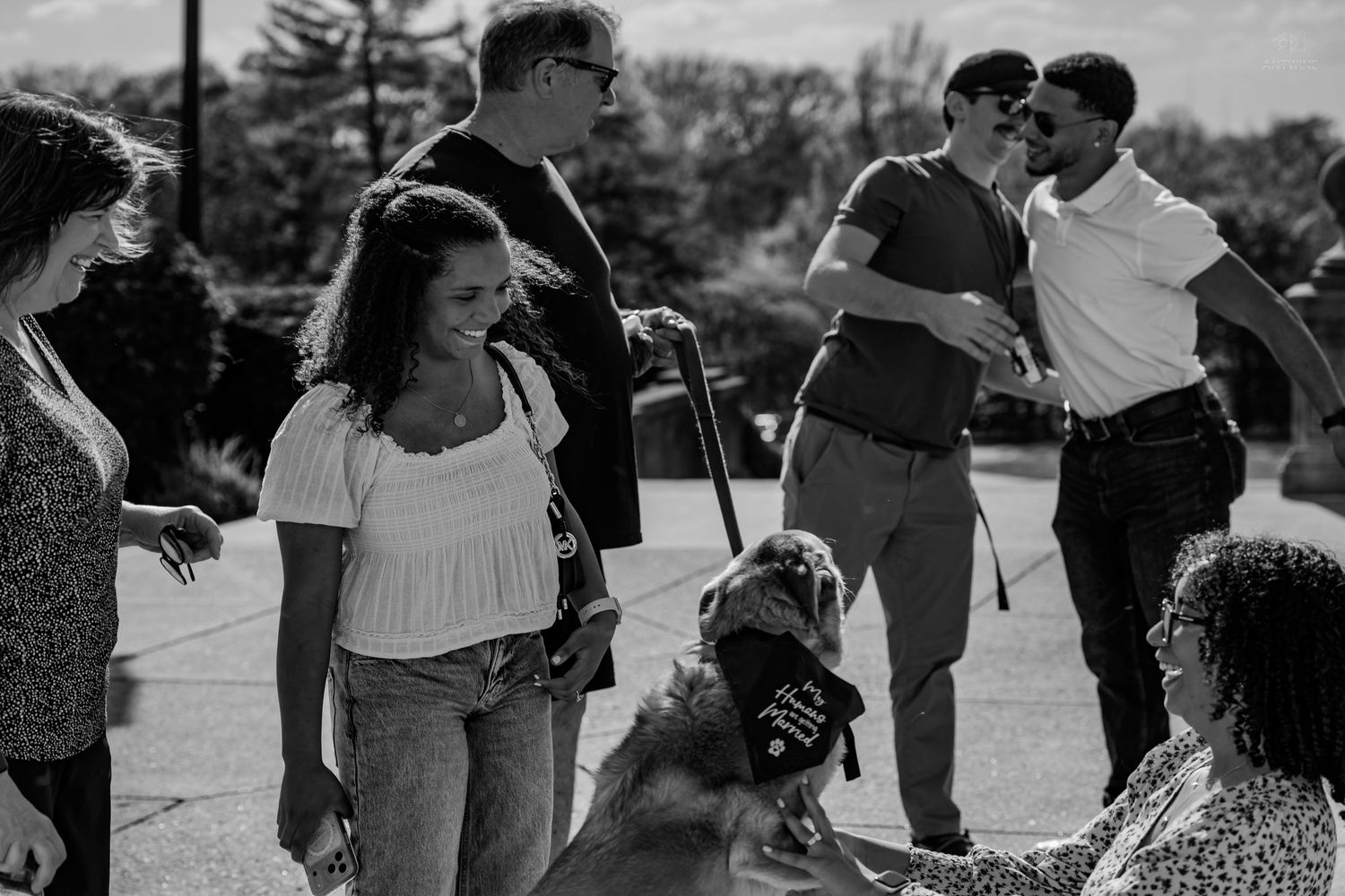 Black and white photo of a group of people gathered outdoors with a dog wearing a bandana.