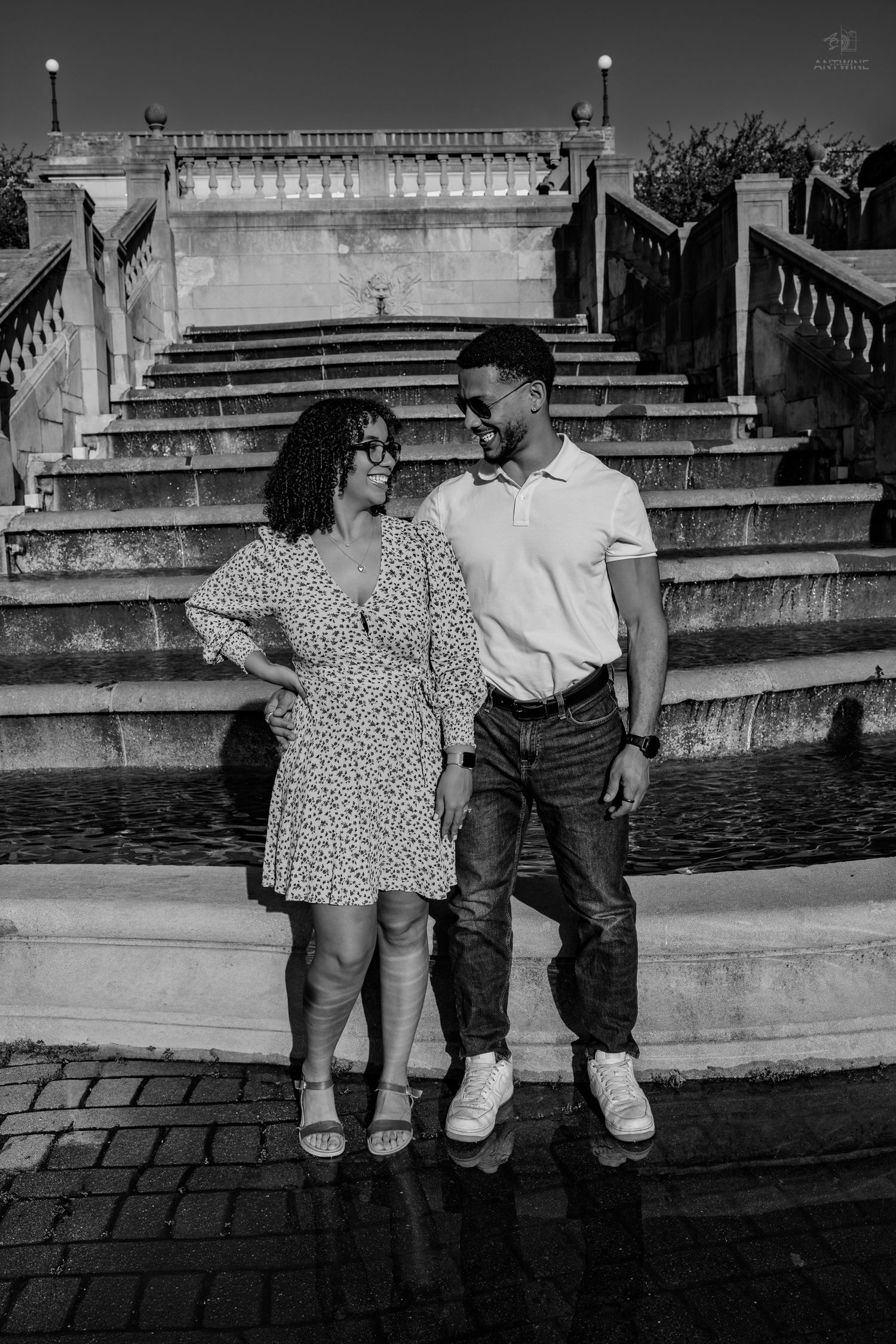 Black and white photo of a couple standing by a fountain with grand staircase in the background.