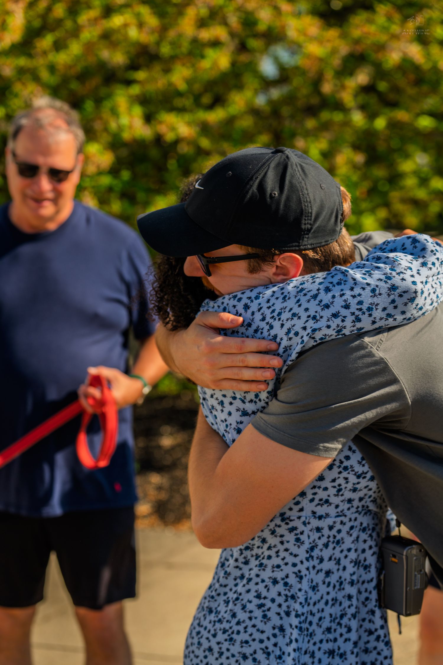Two people share a warm hug outdoors in golden sunlight while a man watches nearby.