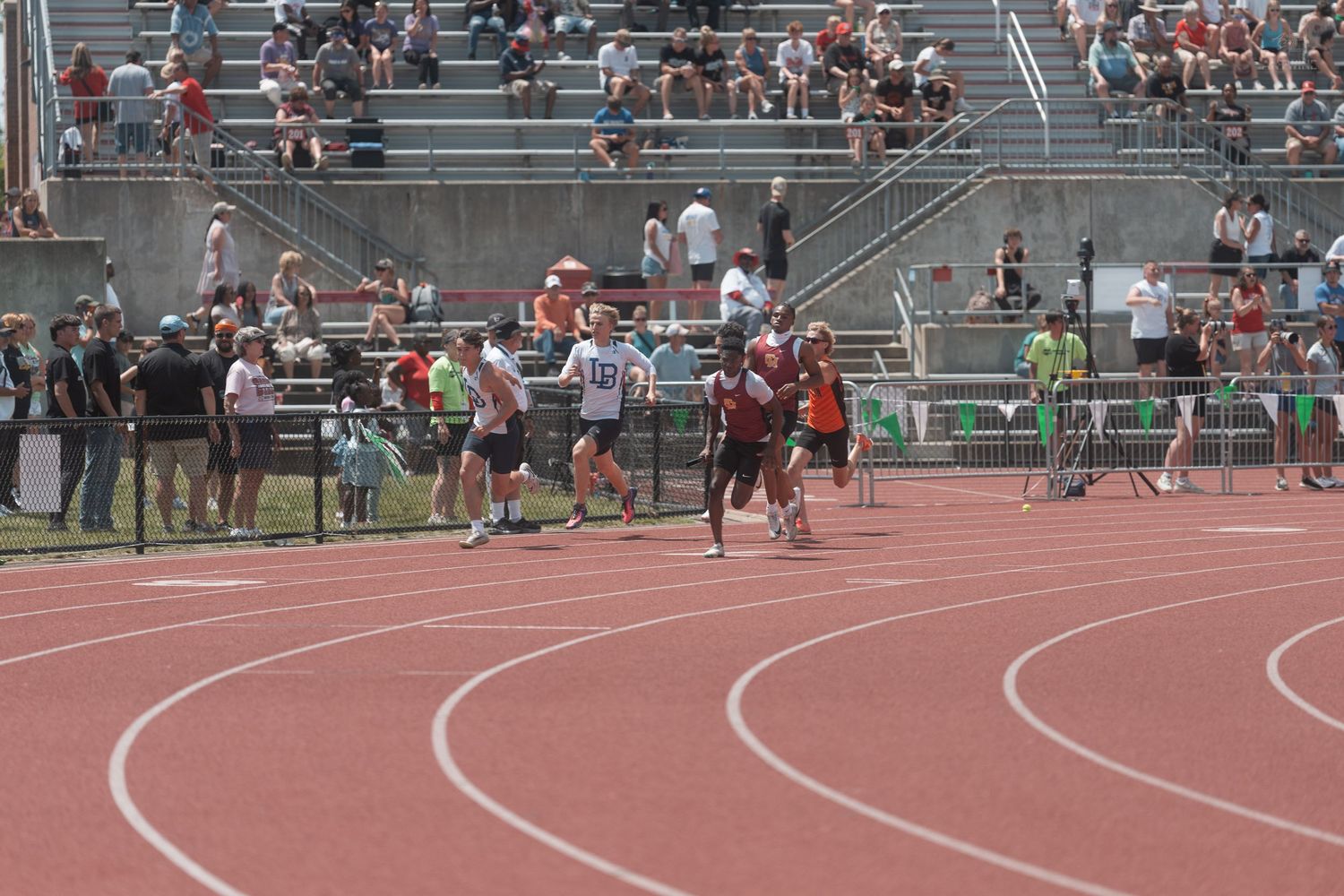 Runners race on a red track at an outdoor athletic meet with spectators filling the stands.