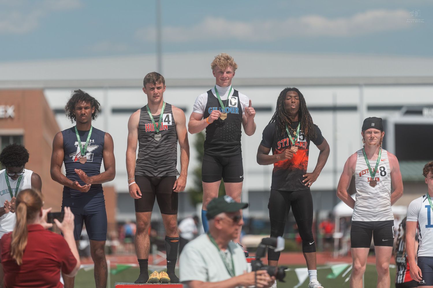 Male high school athletes pose with medals on a numbered awards podium at an OHSAA track meet.