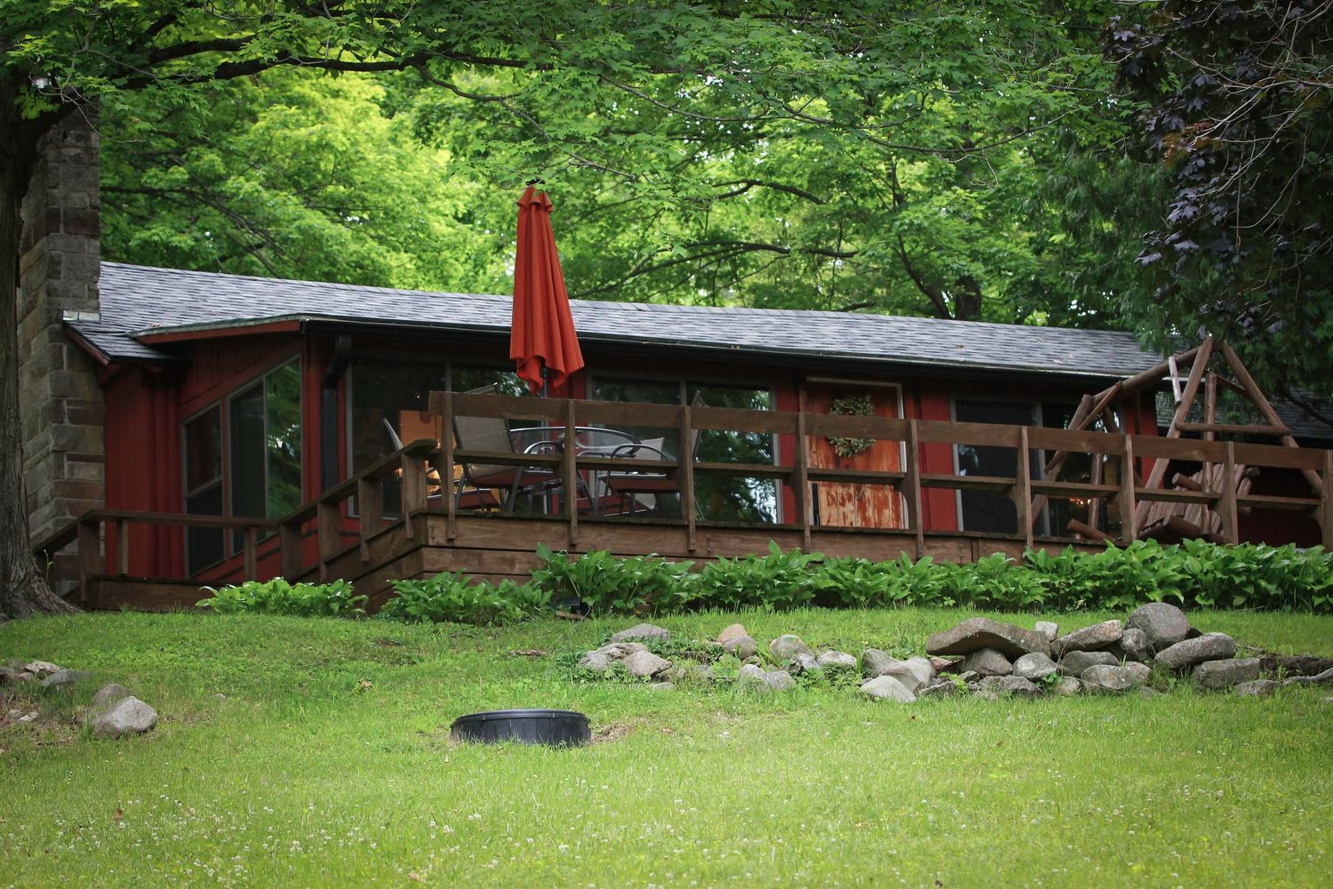 Rustic cabin surrounded by lush green forest with a red patio umbrella and stone landscaping on a grassy lawn.