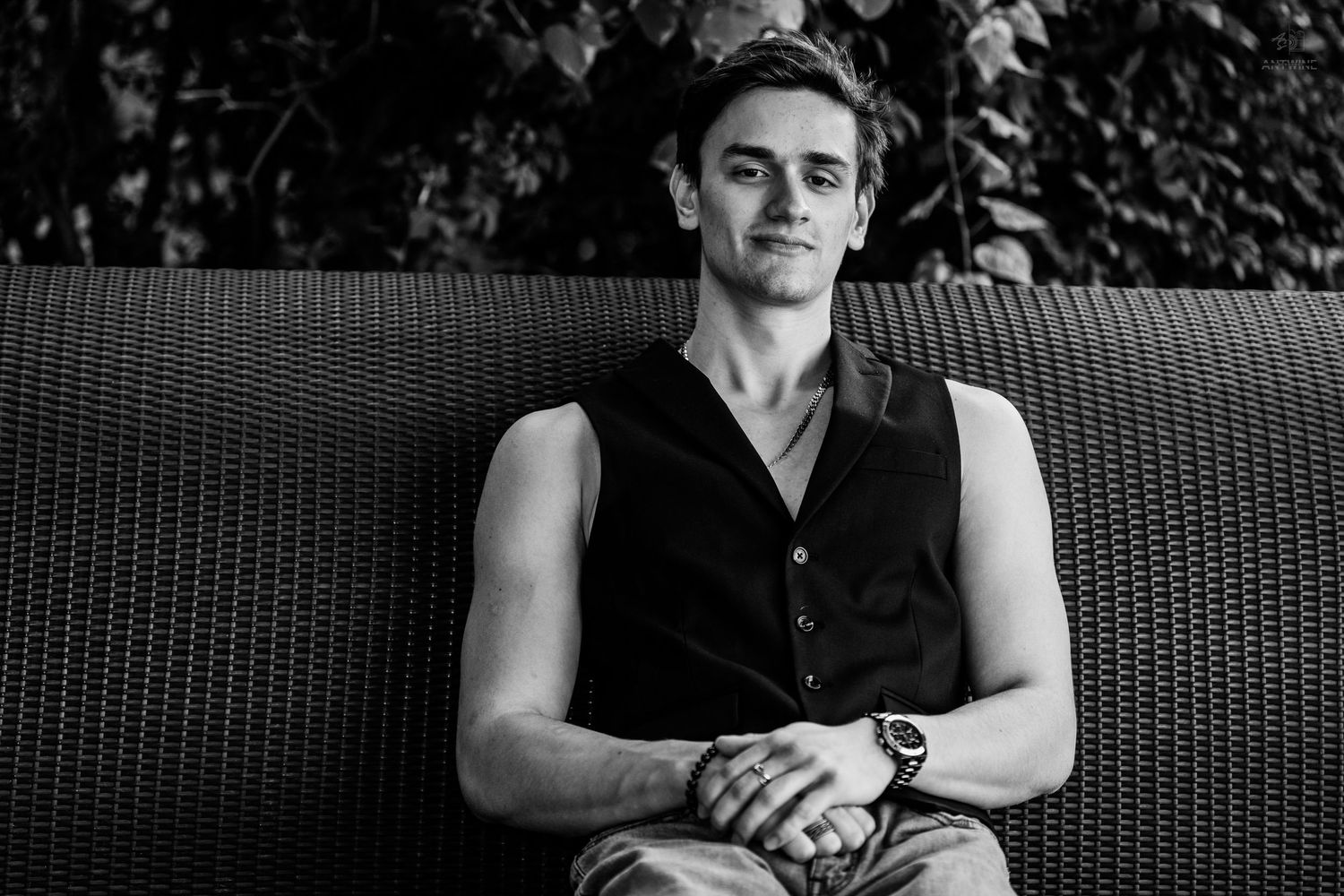 Young man in black sleeveless vest sitting relaxed on wicker bench in black and white photo.