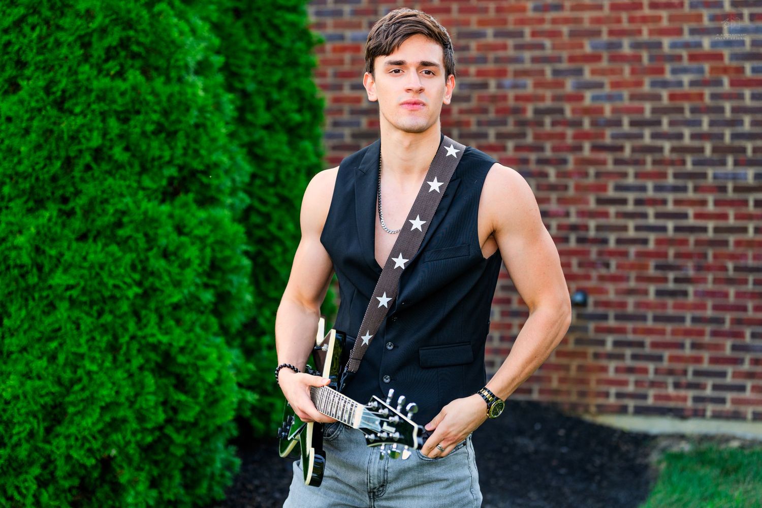 Young male guitarist in black sleeveless star vest holding electric guitar outdoors near brick wall and green tree.