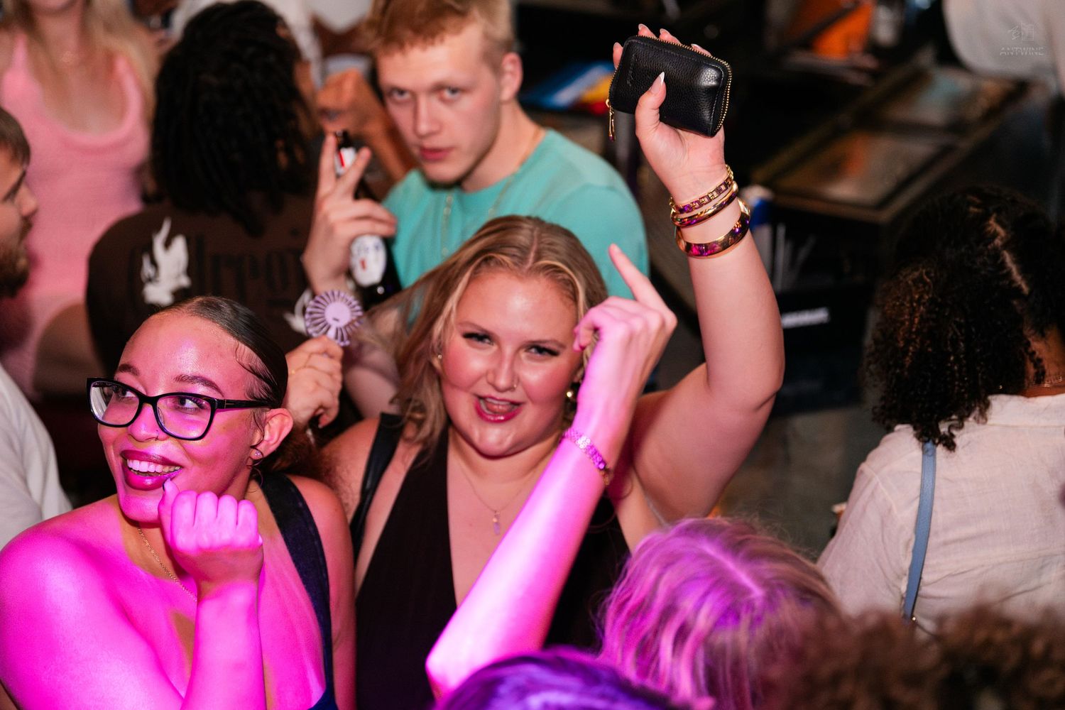 Two women smiling and dancing at a vibrant nightclub party under pink lighting.