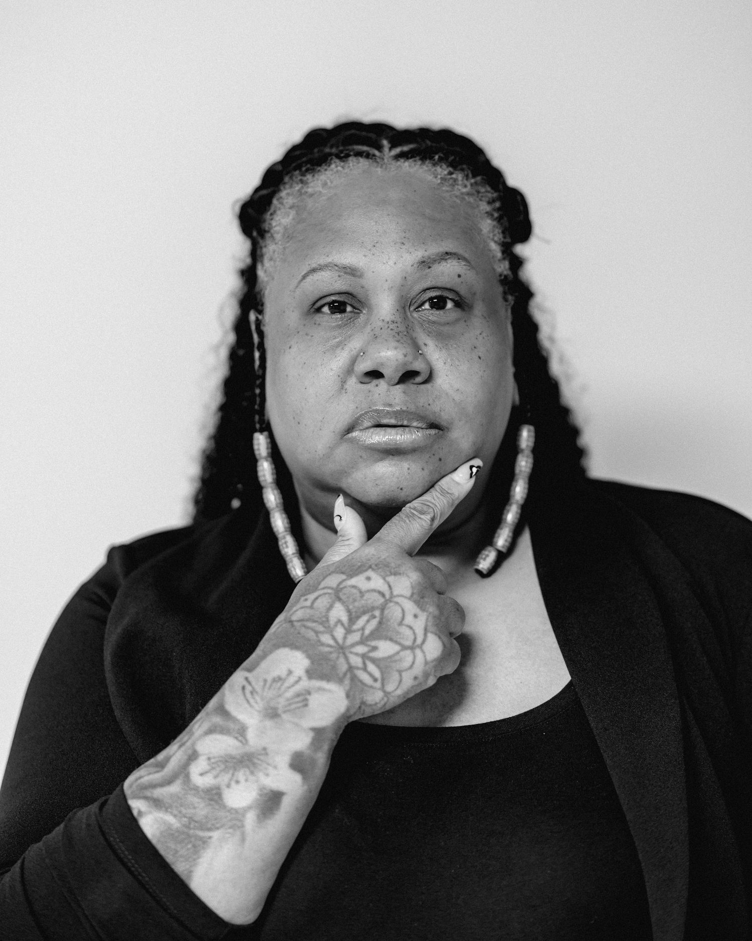Black and white portrait of a woman with braided hair and floral hand tattoo in a thoughtful pose.