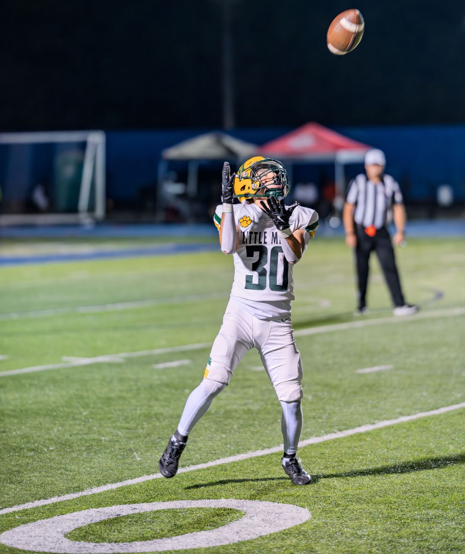 Youth football player #30 in white and gold uniform prepares to catch a football during an evening game.