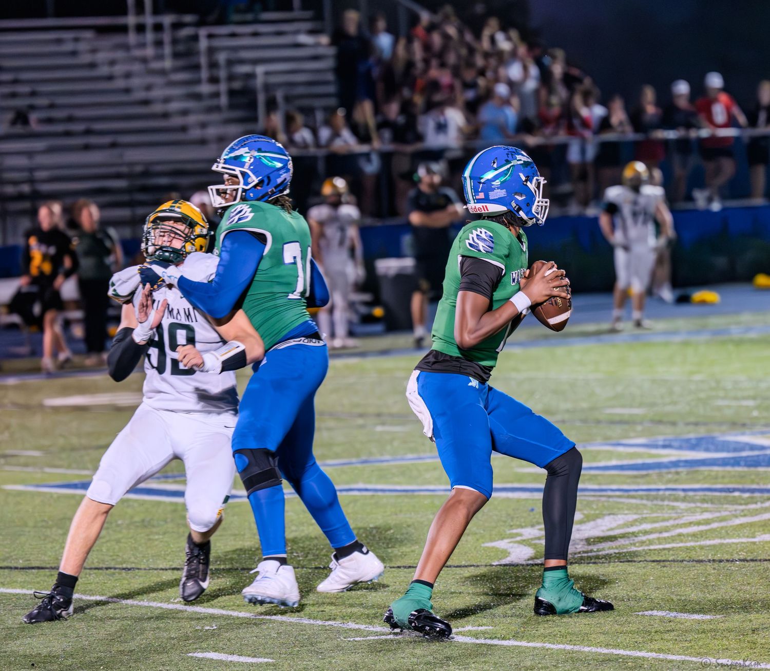 Warriors quarterback drops back to pass while offensive lineman blocks defender during a night football game.