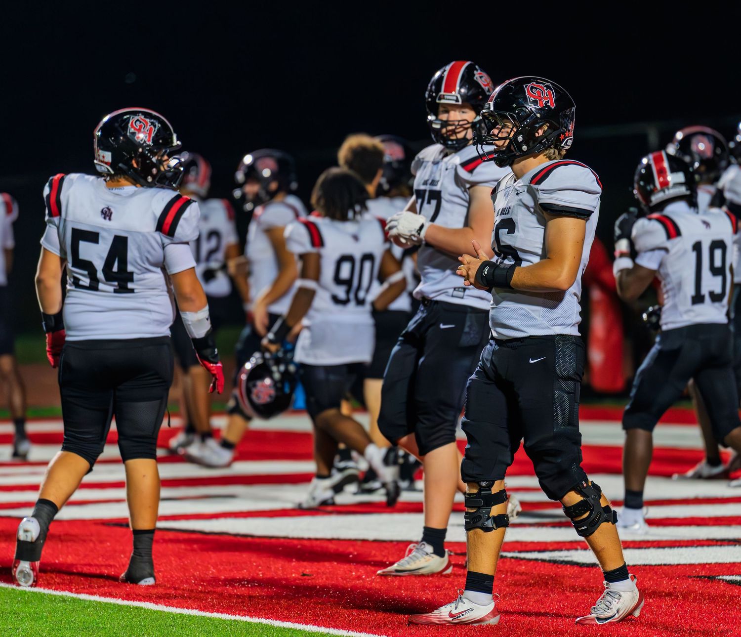 High school football players in white away uniforms gather on the field during a night game.