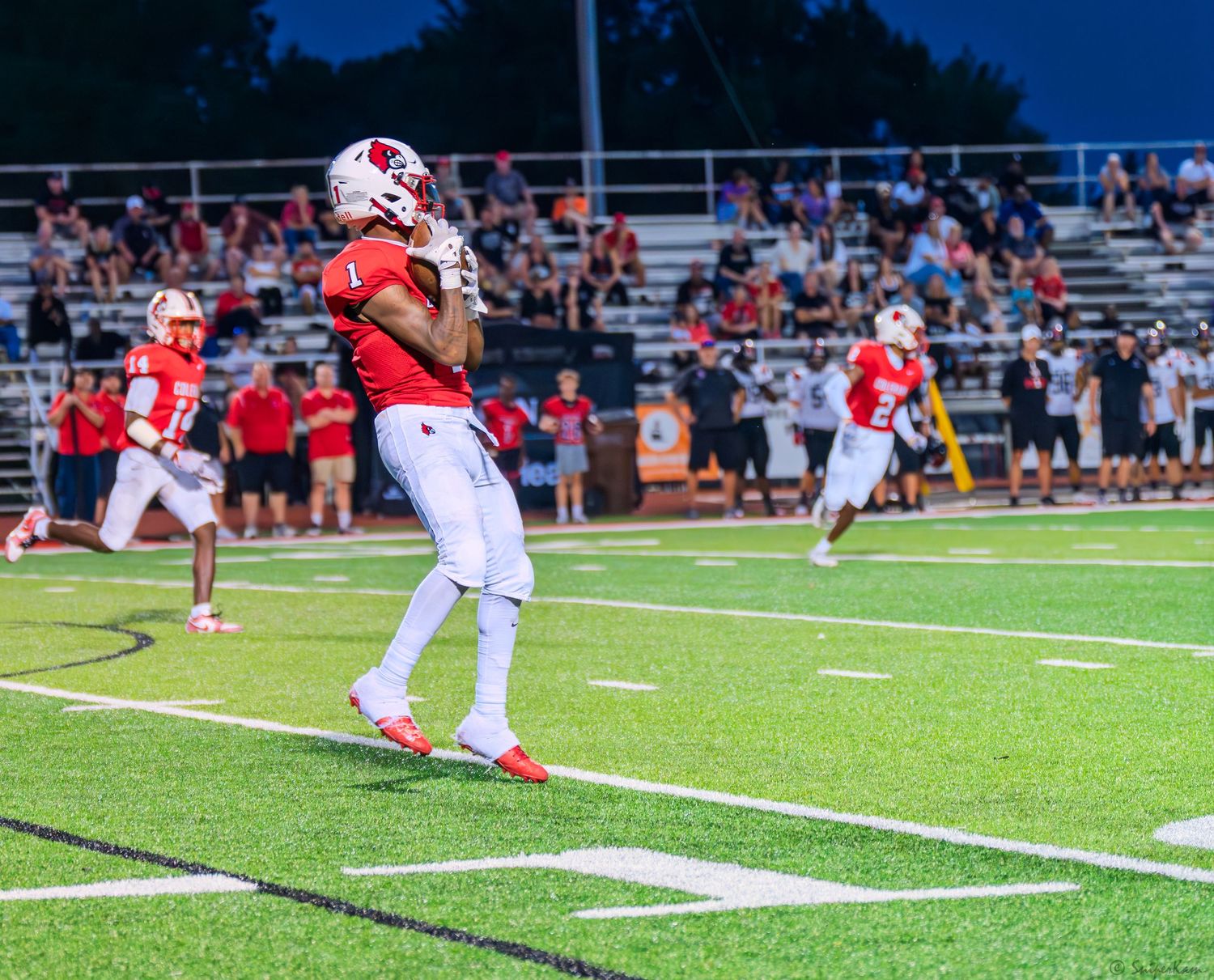 Football player in red uniform celebrates in the end zone after scoring a touchdown at night.