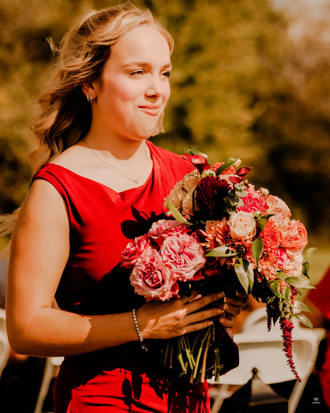 Bridesmaid in red dress holding colorful bouquet of pink and red flowers at outdoor wedding ceremony.
